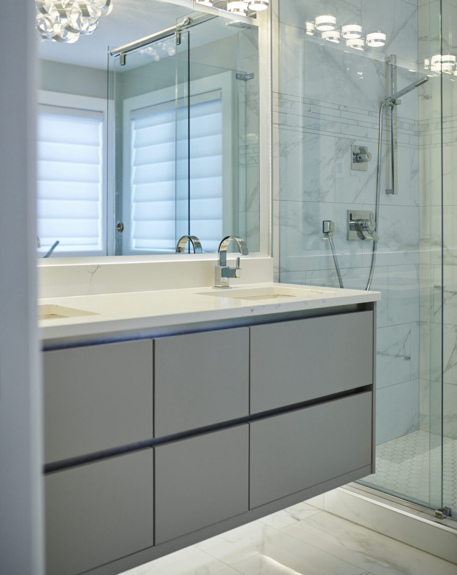 Modern bathroom interior with gray vanity, white countertop, two sinks, and a glass-enclosed shower with marble walls and hexagonal floor tiles.