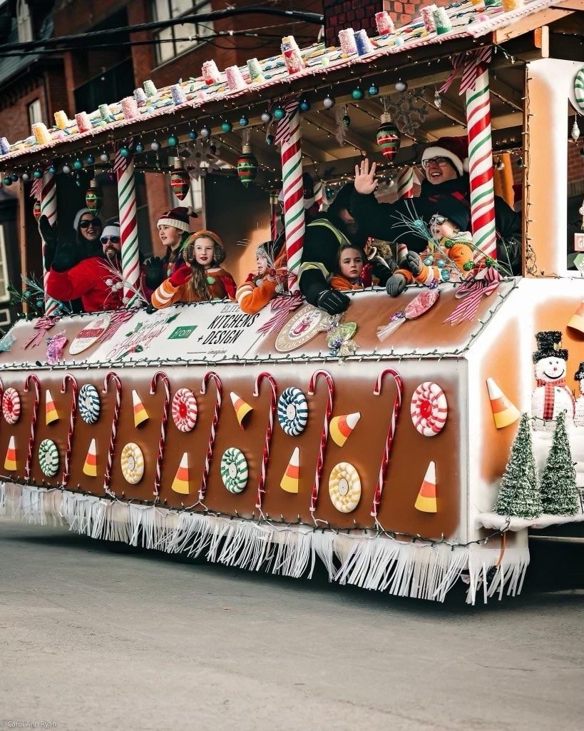 A festive parade float resembling a gingerbread house carries people dressed in holiday attire, waving to onlookers and surrounded by candy-themed decorations.