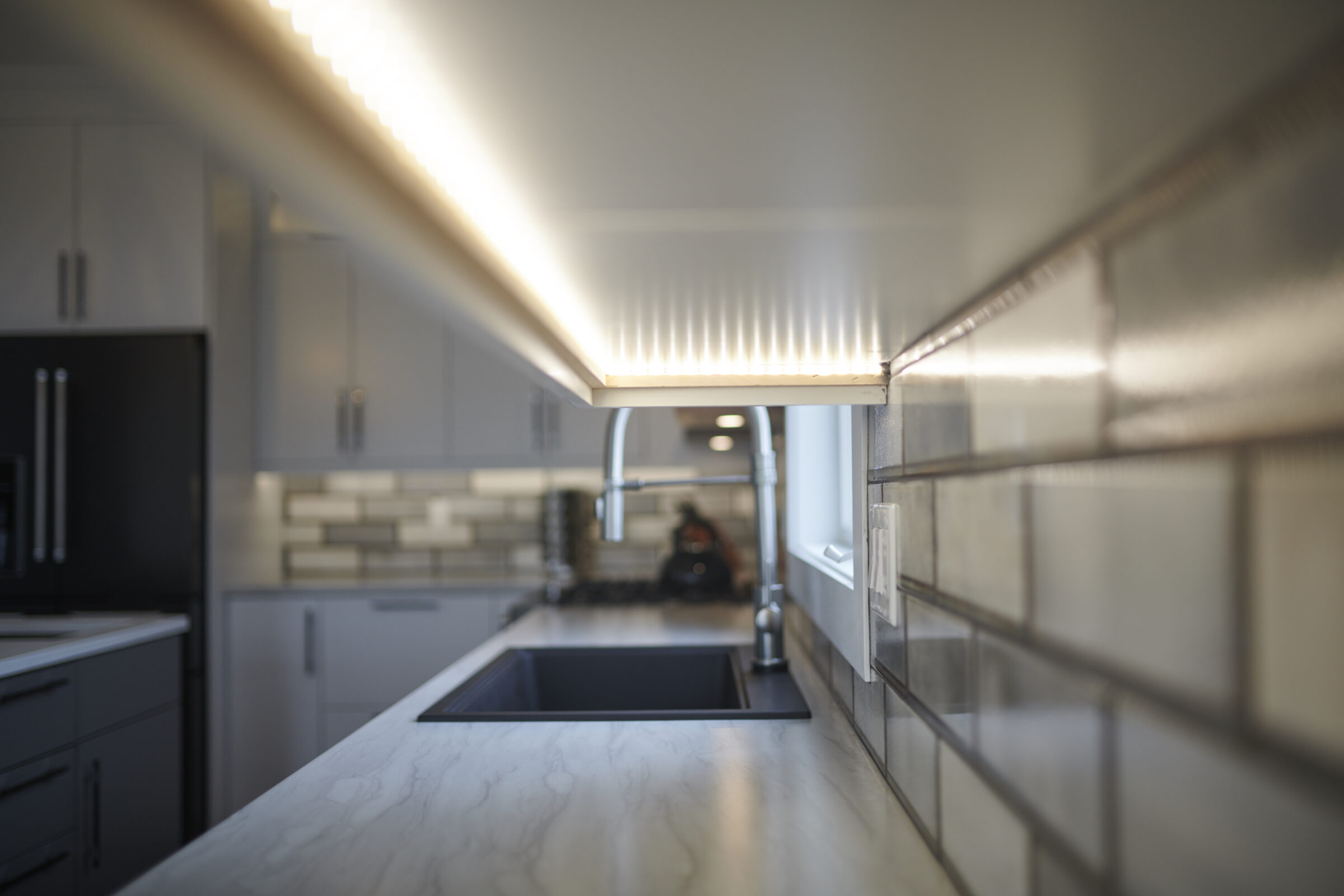 A modern kitchen with under-cabinet lighting, featuring a marble countertop, stainless steel sink, and a blurred background emphasizing the illuminated space.
