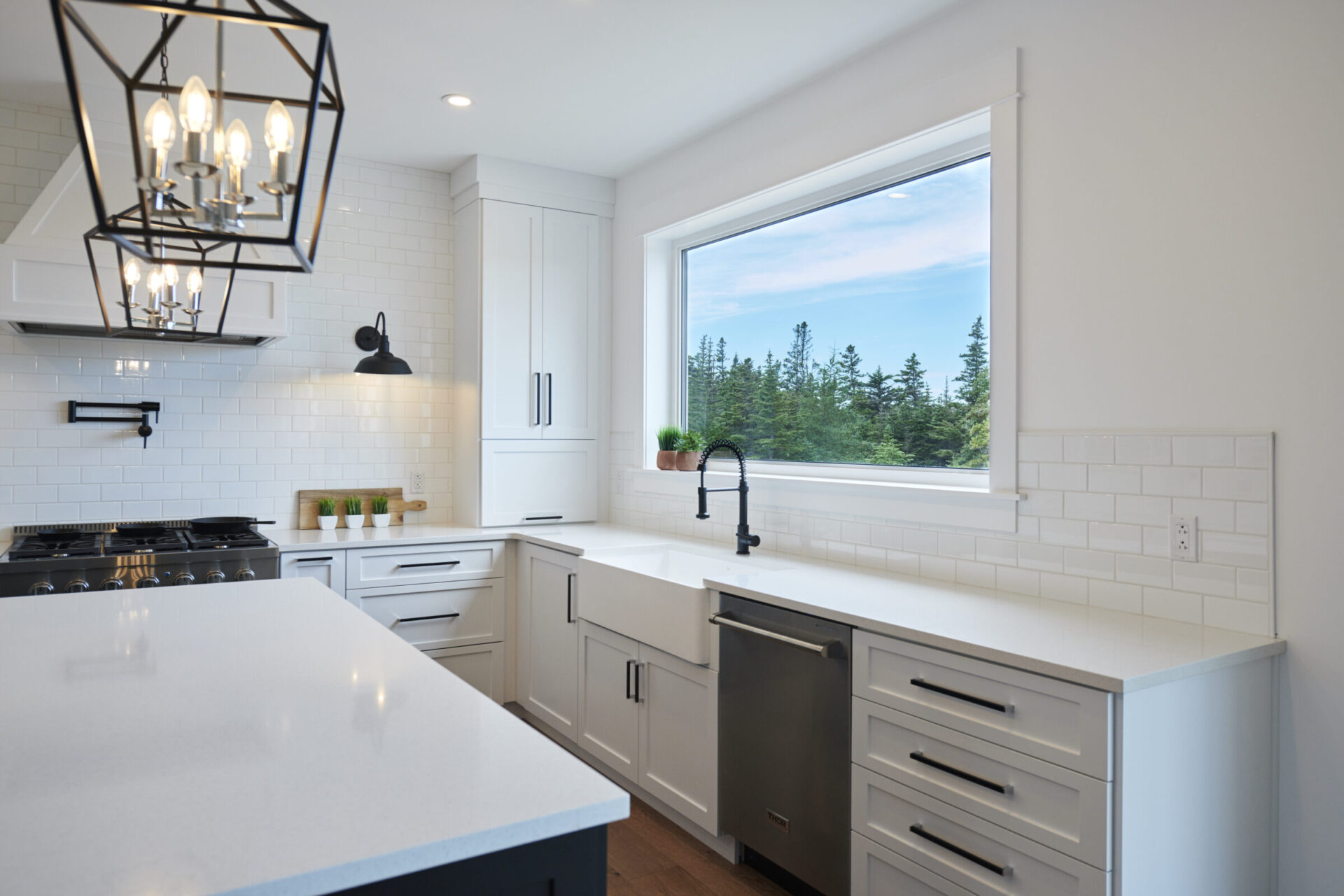 Modern kitchen interior with white cabinets, subway tiles, and stainless steel appliances. A window shows a forested view. Chic pendant light hangs above.