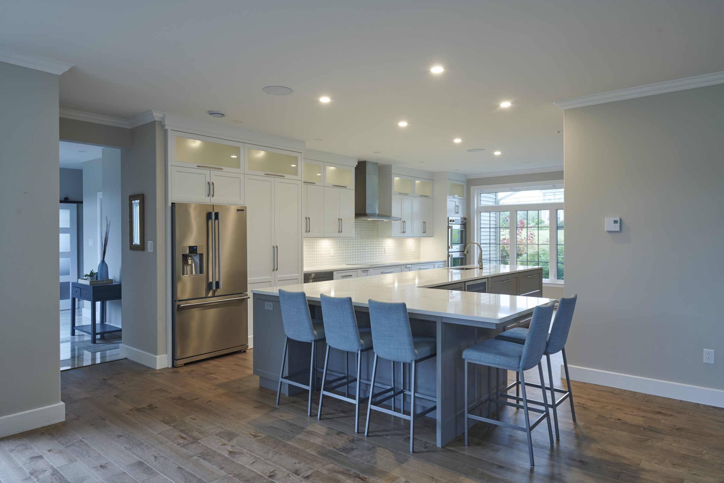 A spacious, modern kitchen with white cabinetry, stainless steel appliances, a large island with blue stools, and recessed lighting in a clean design.