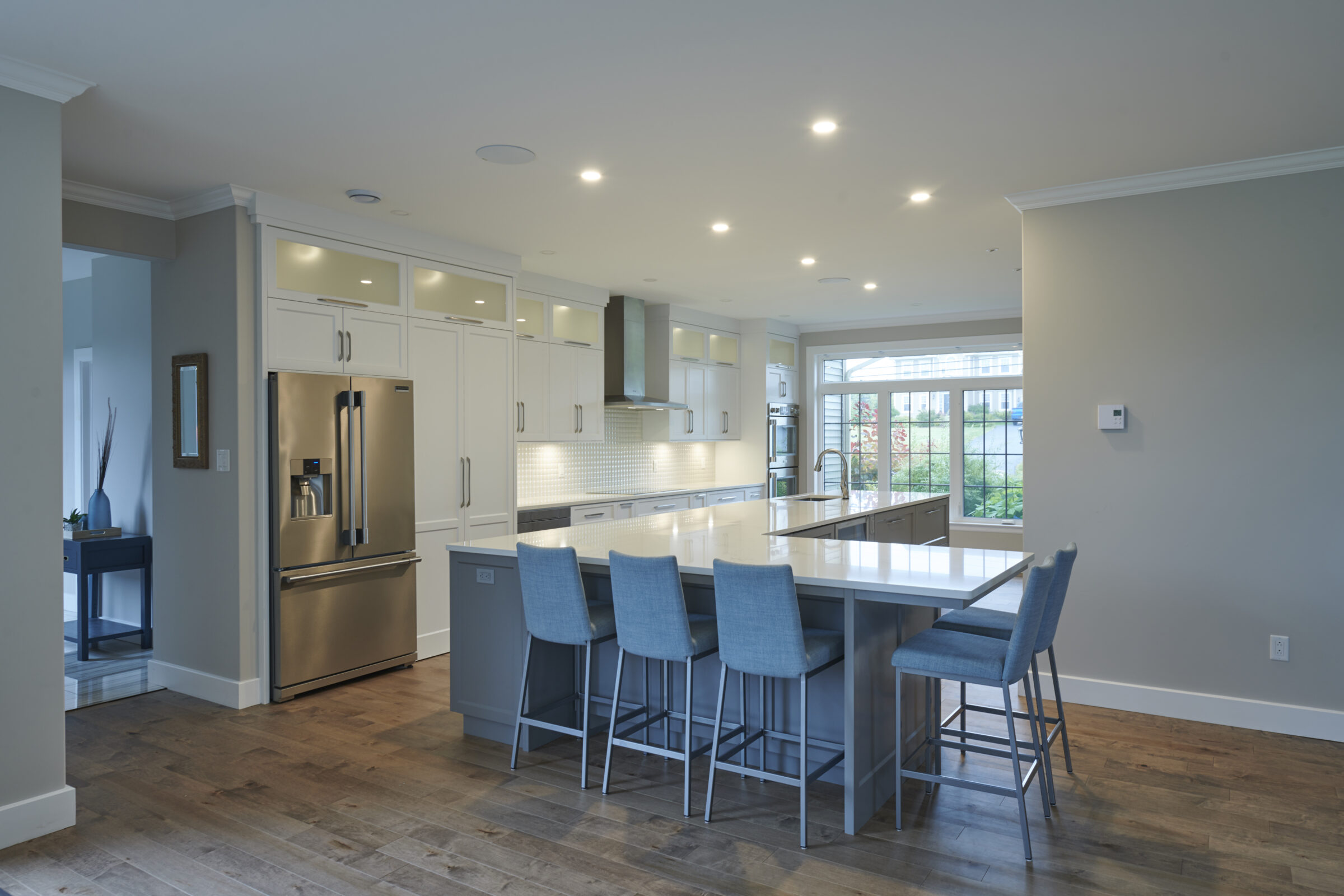 A modern kitchen with white cabinetry, stainless steel appliances, and a central island with blue stools. Brightly lit with large windows and recessed lighting.