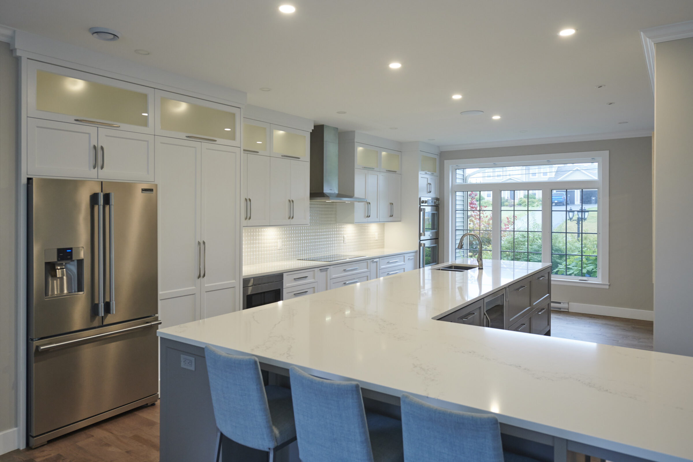 Modern kitchen interior with white cabinetry, stainless steel appliances, marble countertops, and blue chairs. Recessed lighting and a garden view through the window.