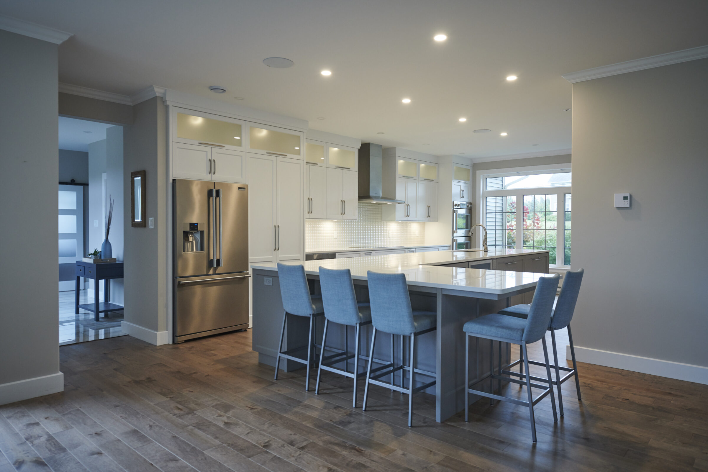 A modern kitchen with white cabinetry, stainless steel appliances, a central island with blue stools, hardwood floors, and recessed ceiling lights.