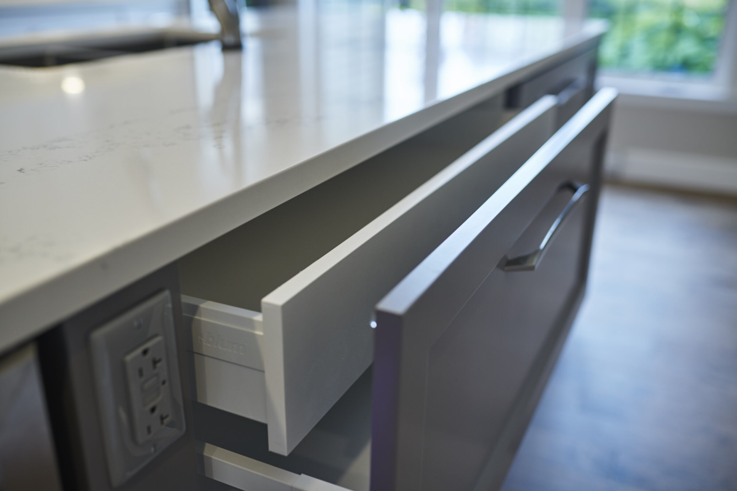 An open modern kitchen drawer with a silver handle, integrated power outlets inside, showcasing tidy storage solutions, and a view of a window.