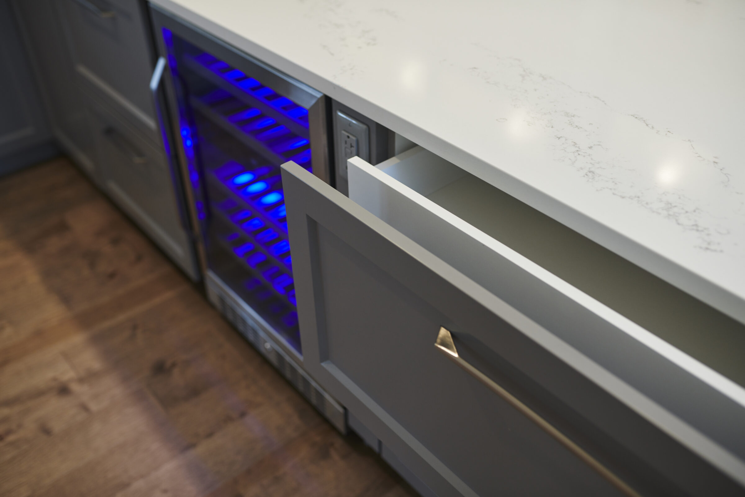 A modern kitchen with open drawers featuring a glowing blue light, sleek handles, a marble countertop, and a hardwood floor. The design is contemporary.