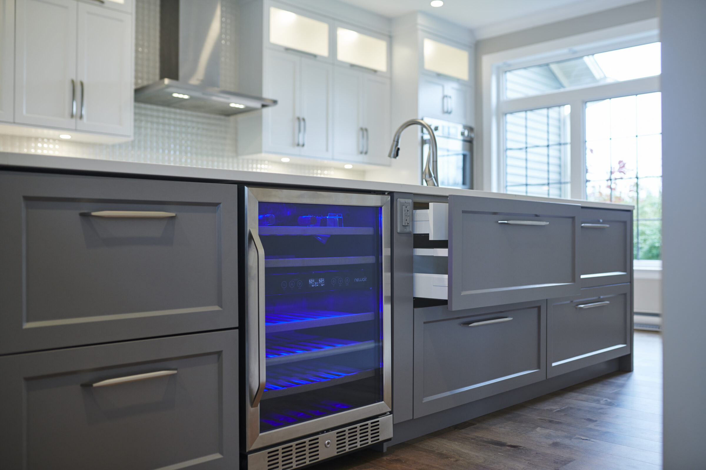 Modern kitchen interior showing gray cabinets, a wine fridge with blue lighting, white countertops, and a stainless steel sink near a bright window.
