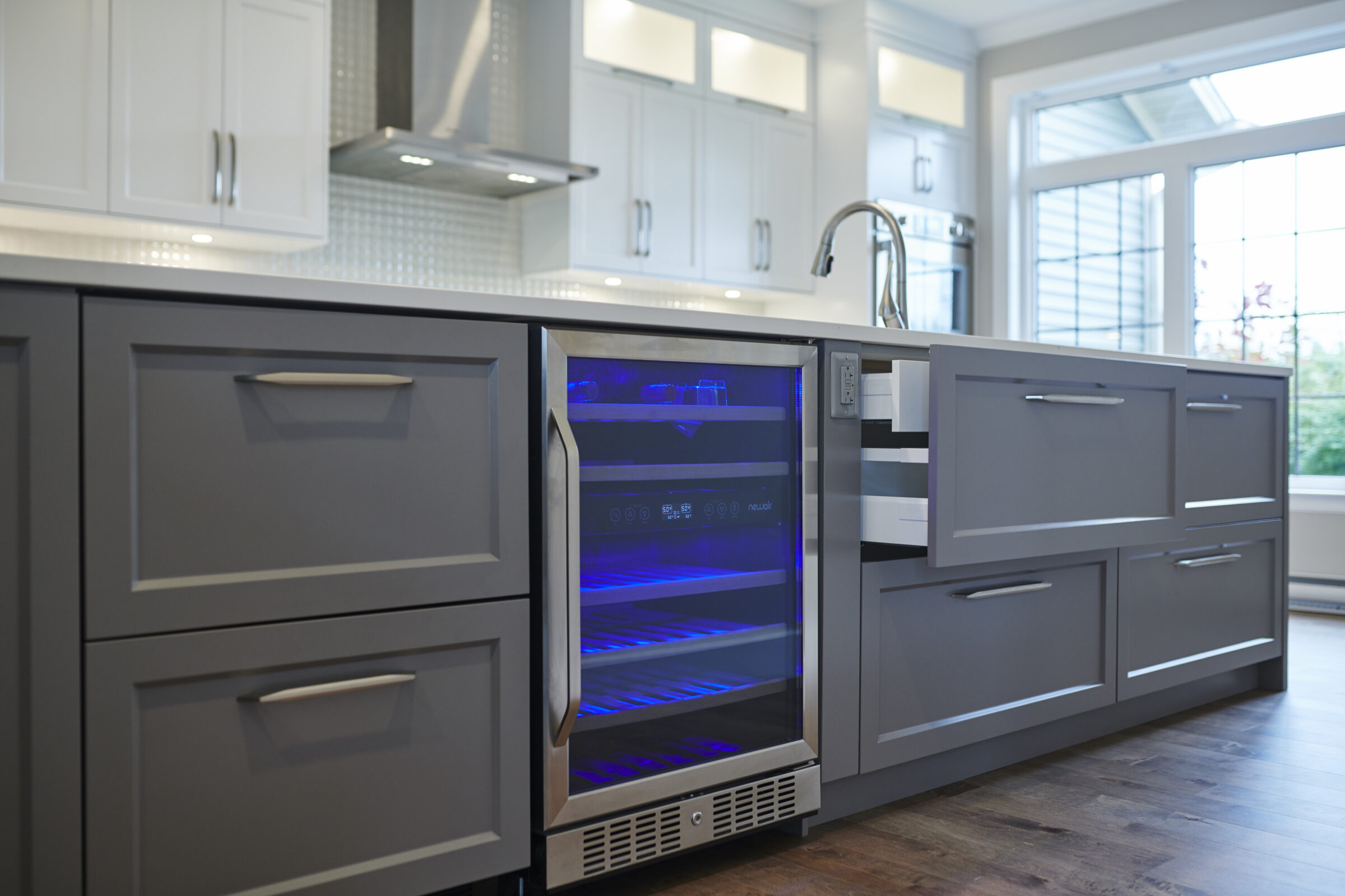 A modern kitchen with white and gray cabinetry, a wine fridge with blue lighting, stainless steel appliances, and a window letting in natural light.