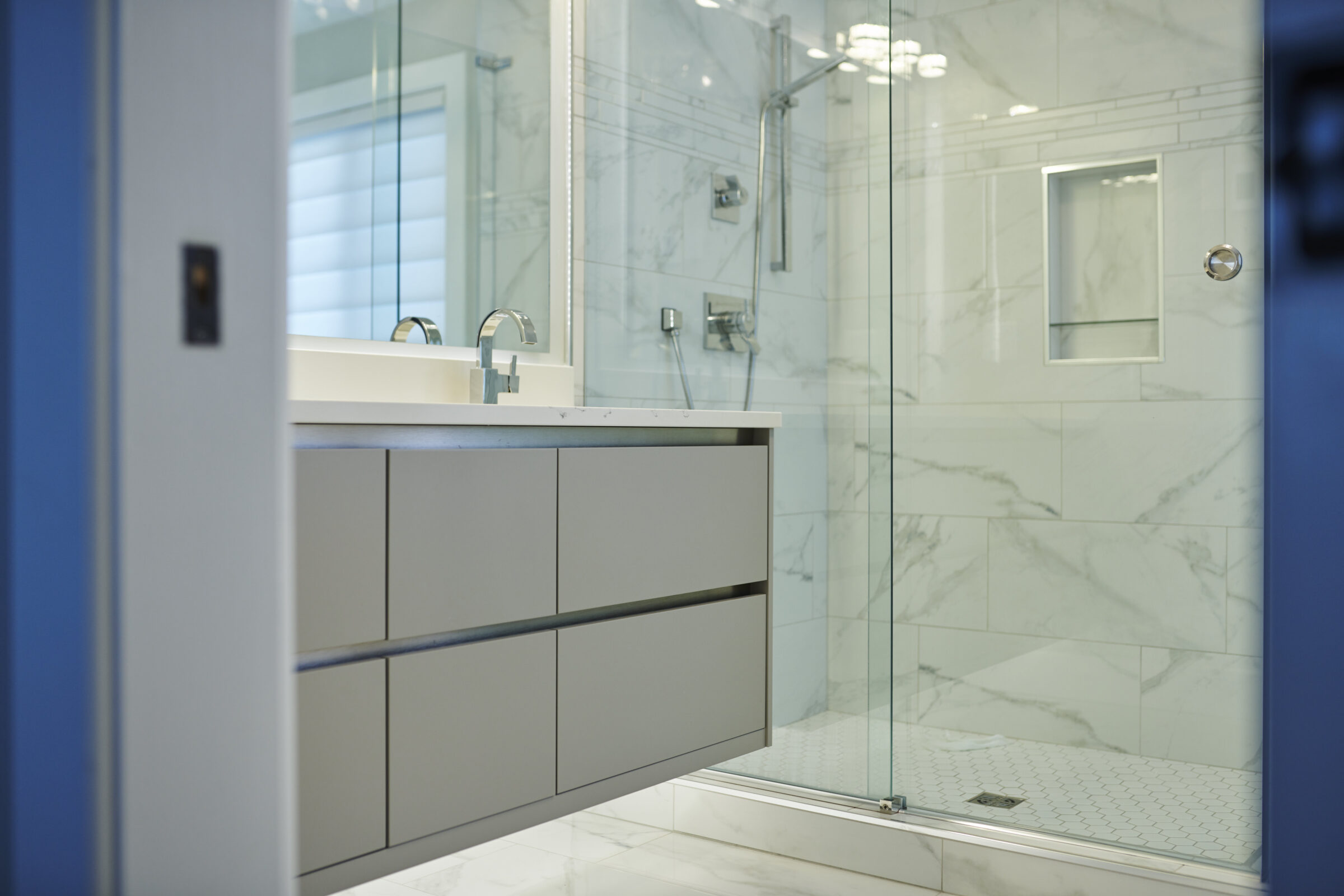 Modern bathroom with marble tiles, featuring a frameless glass shower, grey vanity cabinet, and a blurred reflection of a faucet in the mirror.