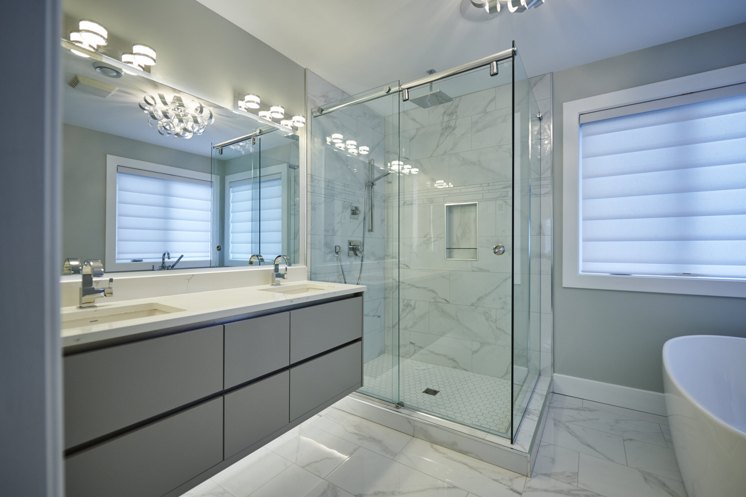 Modern bathroom with marble tiles, glass shower, double vanity, large mirror, freestanding tub, elegant lighting fixtures, and a window with shades.