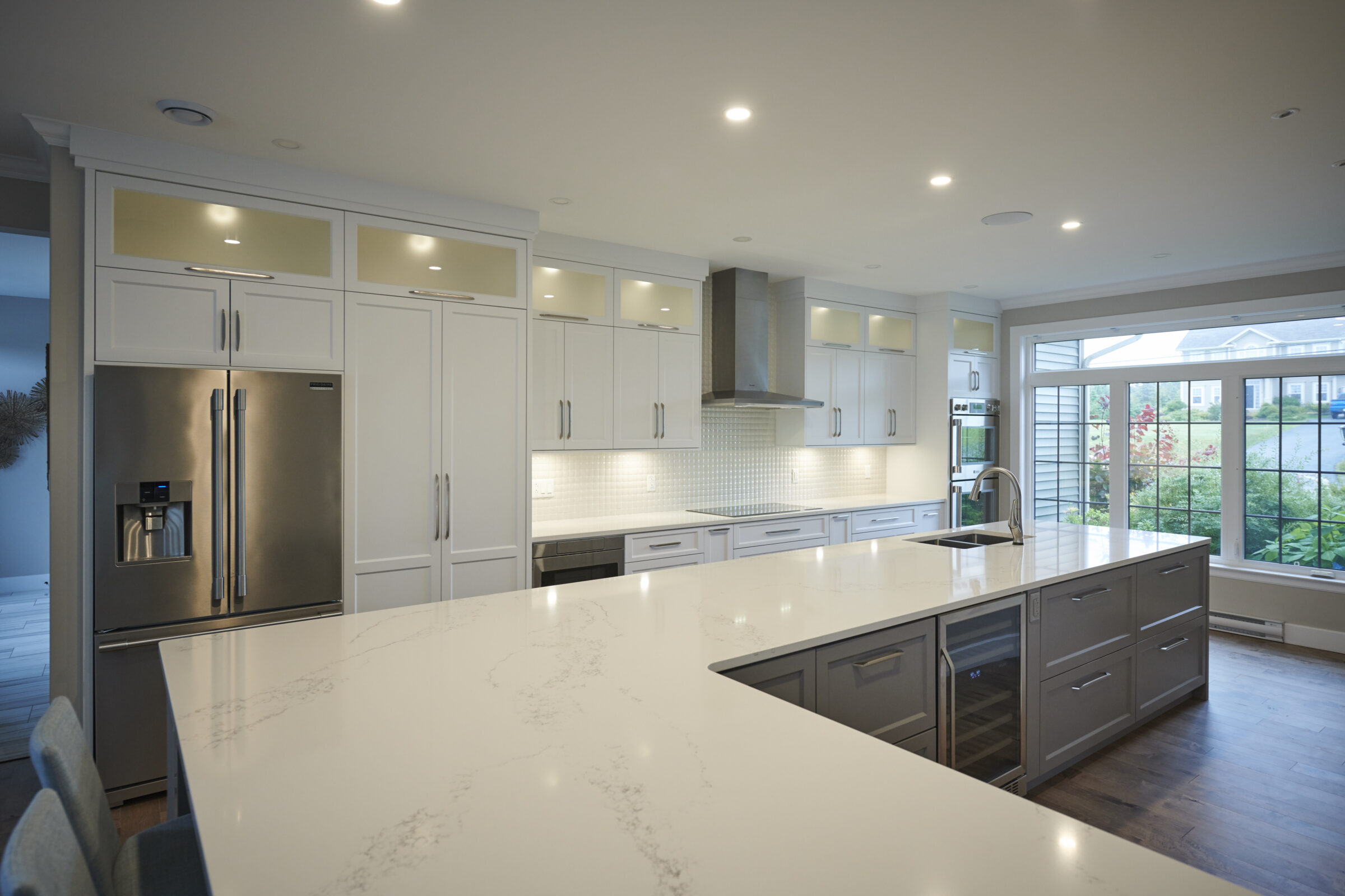 Modern kitchen with white cabinetry, stainless steel appliances, marble countertops, recessed lighting, and a view to an outdoor garden through large windows.