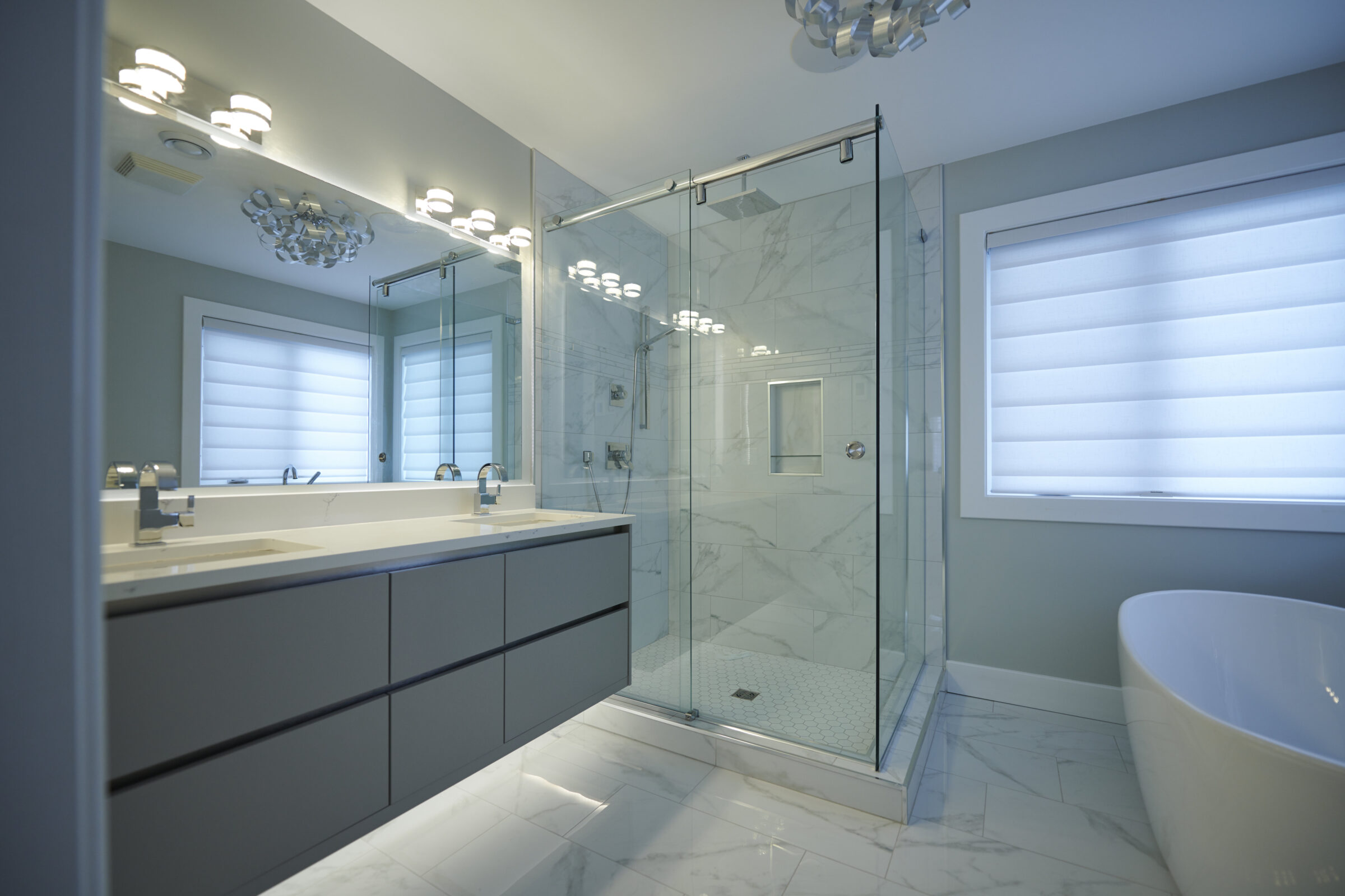 A modern bathroom with a large mirror, dual-sink vanity, glass-enclosed shower, freestanding tub, marble tiles, and bright lighting fixtures.