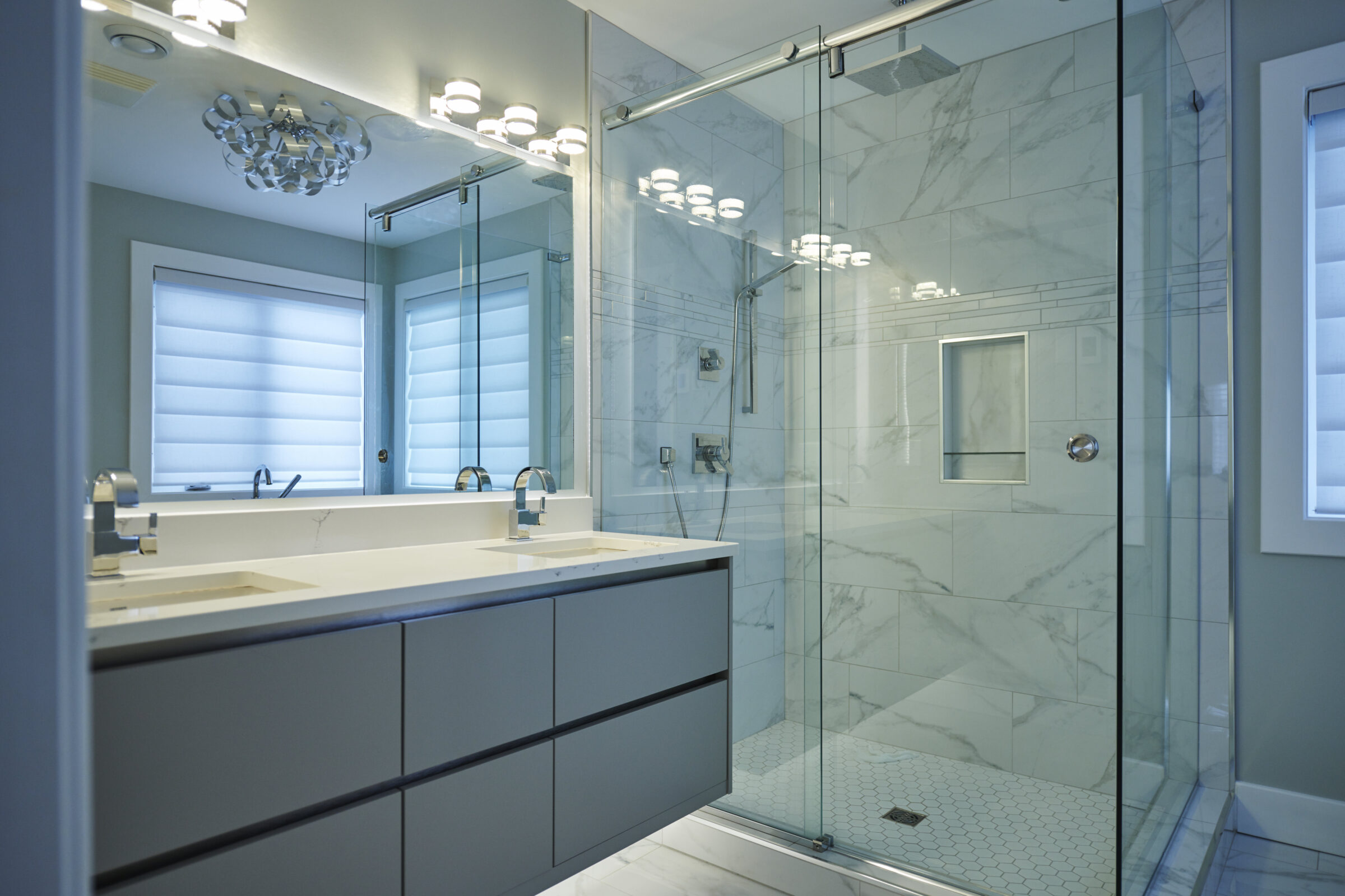 Modern bathroom featuring a dual vanity with a large mirror, sconce lighting above, and a glass-enclosed shower with marble tiles throughout.