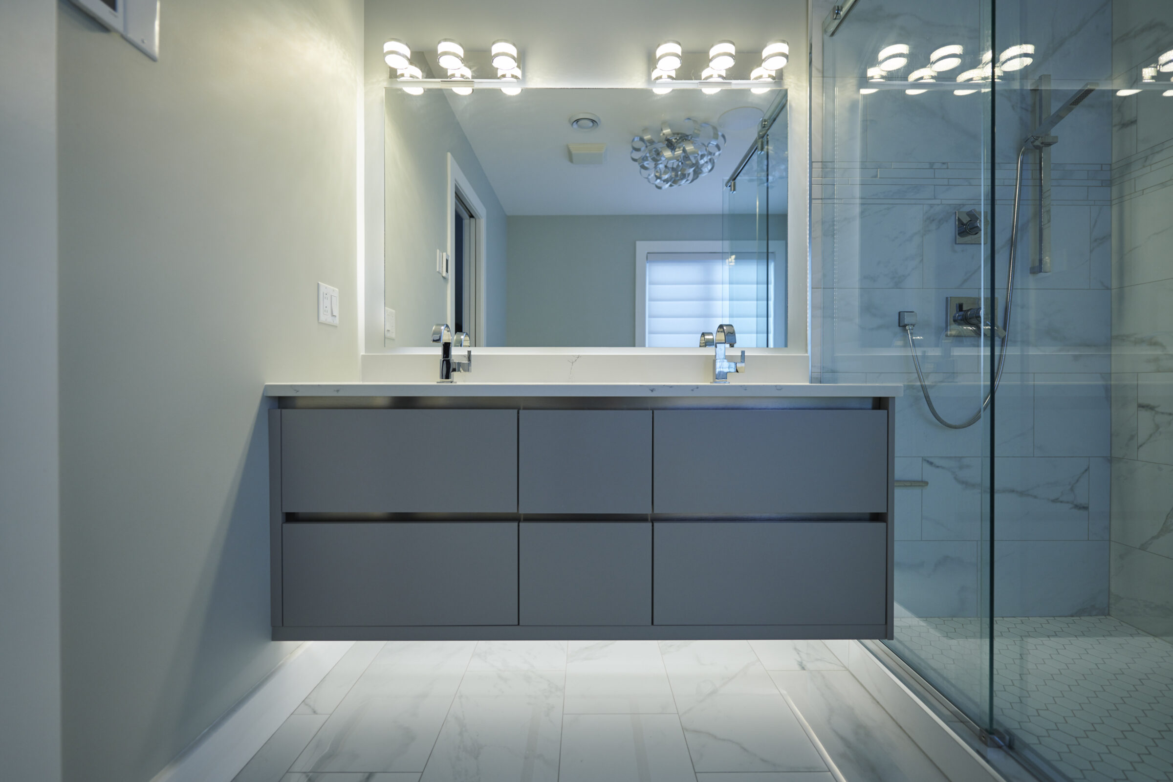 A modern bathroom with marble walls, a floating vanity with dual sinks, a large mirror, overhead lights, and a glass-enclosed shower area.