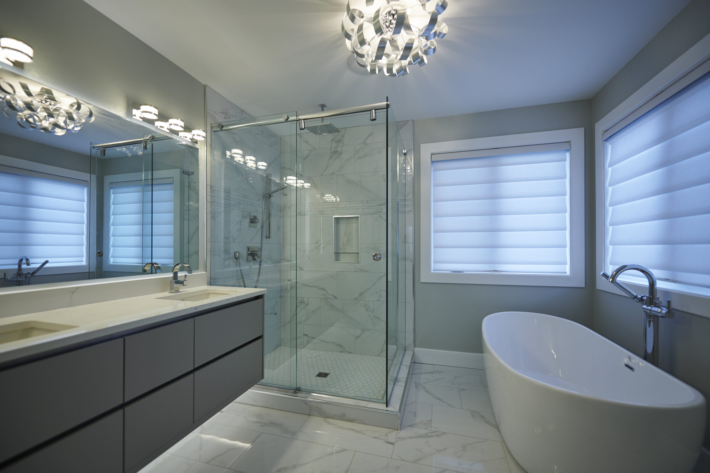 A modern bathroom with a glass shower stall, freestanding tub, double vanity, decorative lighting, and window shades in a cool color scheme.