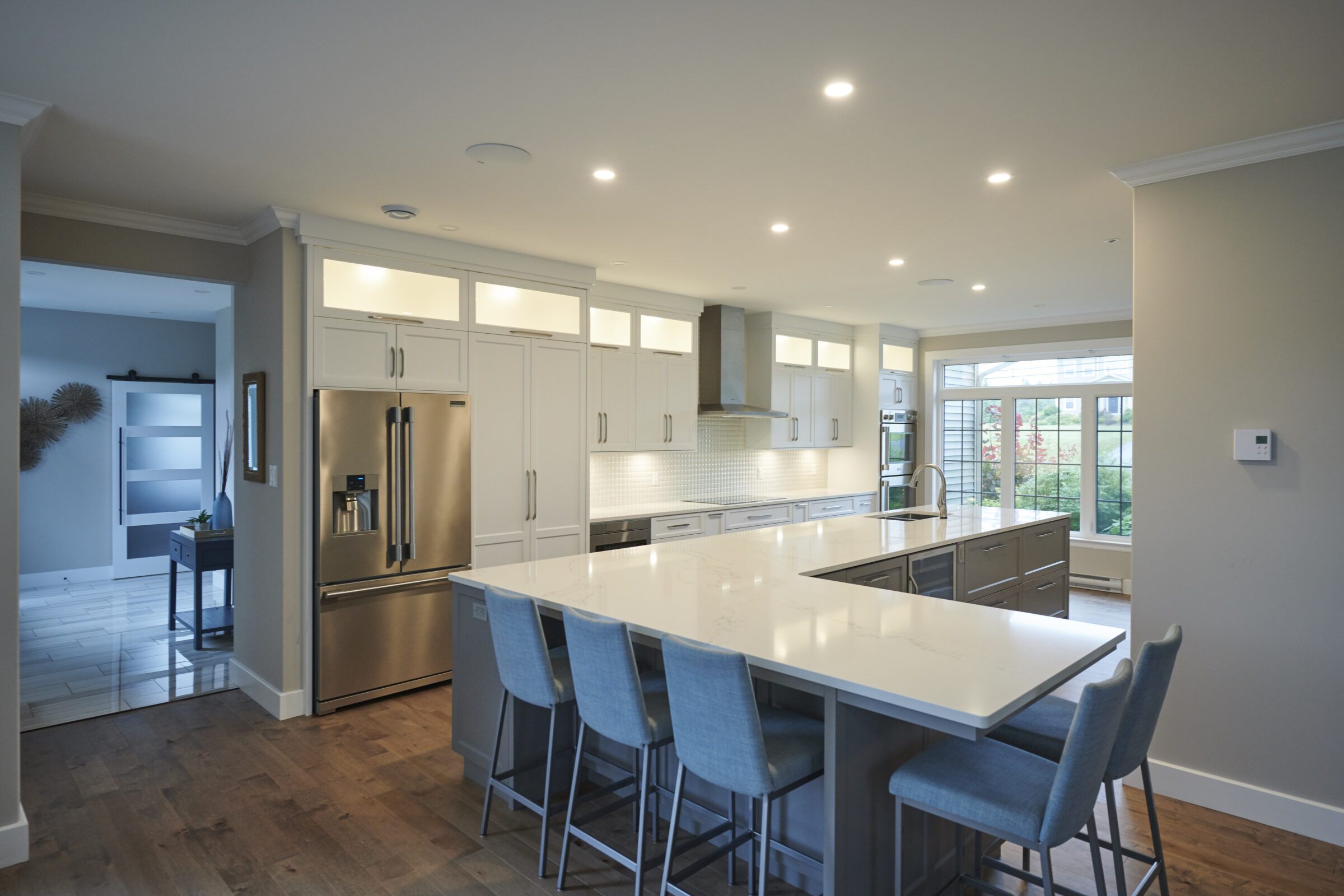 A modern kitchen with stainless steel appliances, white cabinetry, recessed lighting, and a central island with blue stools. There's hardwood flooring and a garden view.