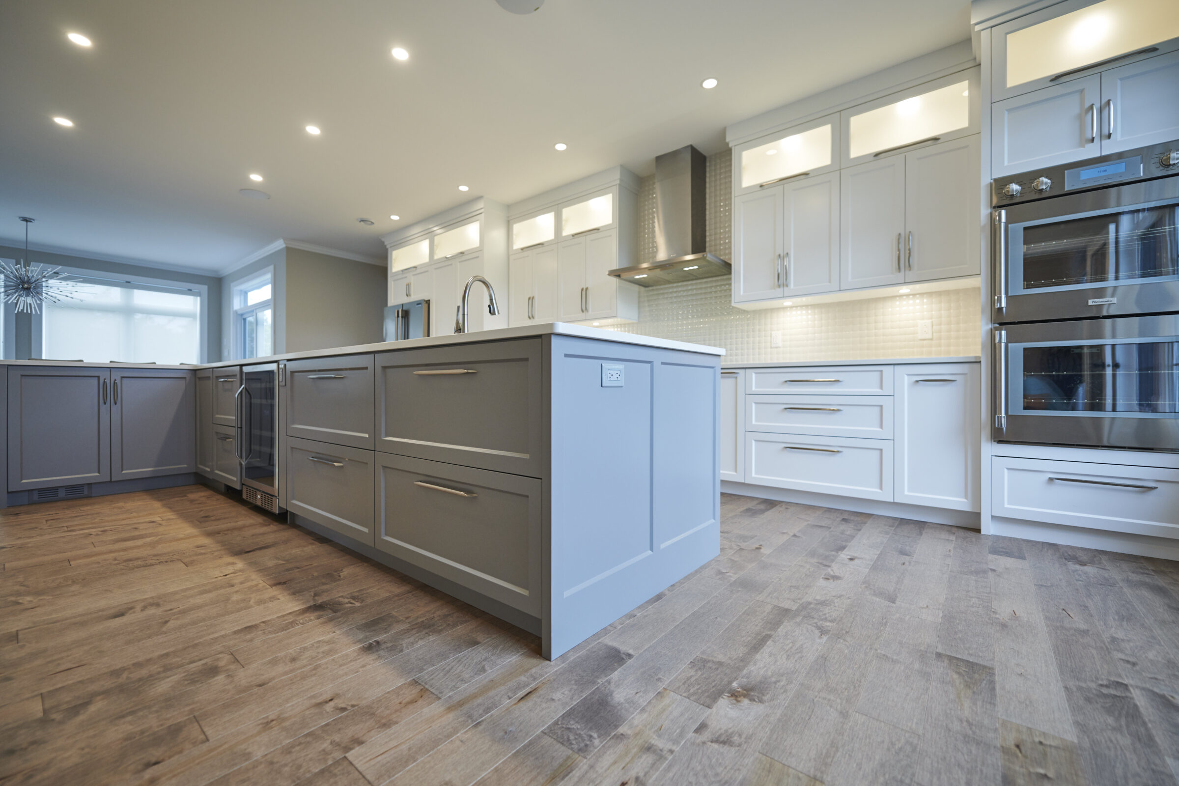 The image shows a modern kitchen interior with white cabinets, stainless steel appliances, gray island, hardwood floors, and recessed lighting. No people are visible.
