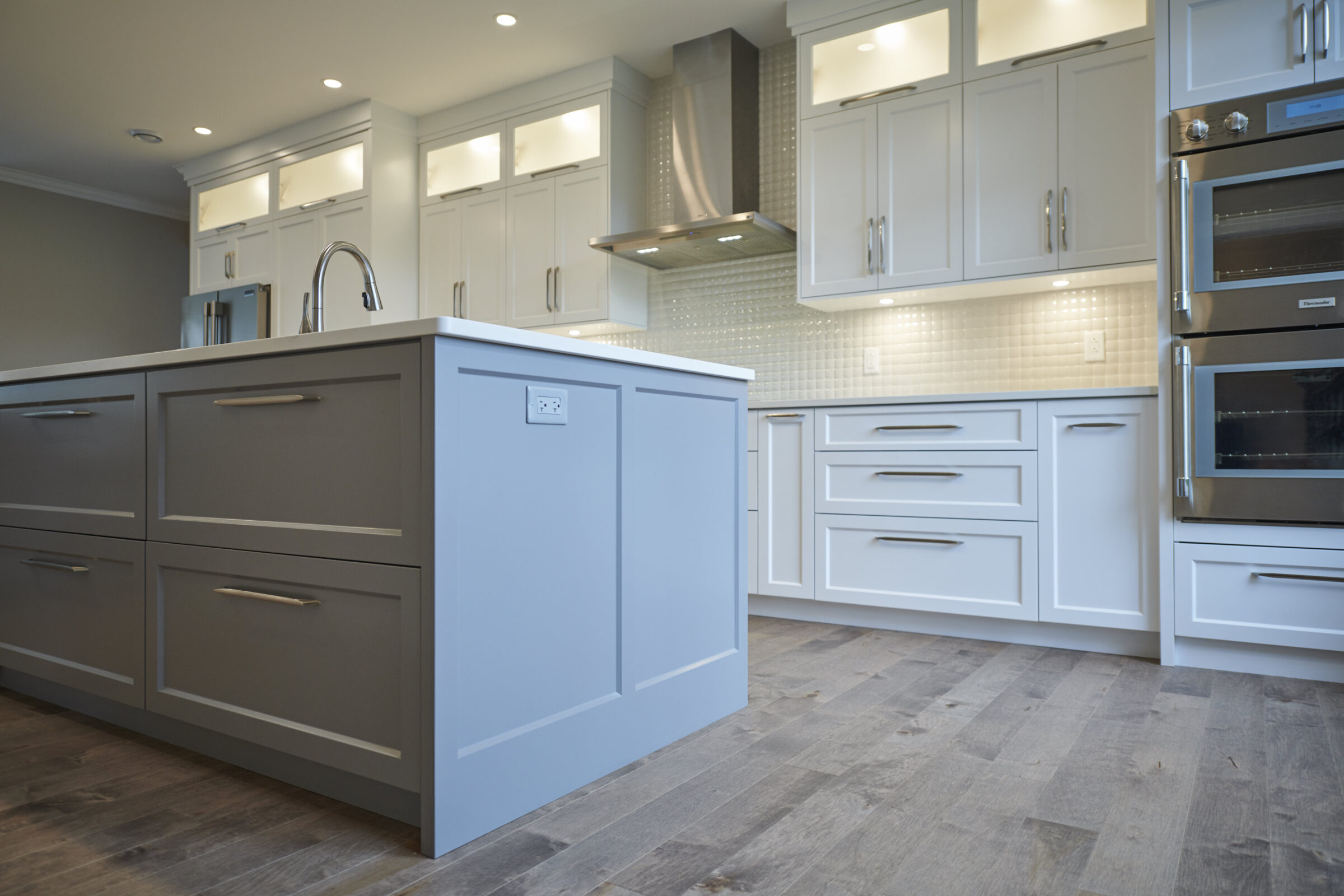 Modern kitchen interior with white cabinetry, gray island, stainless steel appliances, herringbone tile backsplash, and hardwood flooring. Bright, clean, and contemporary design.