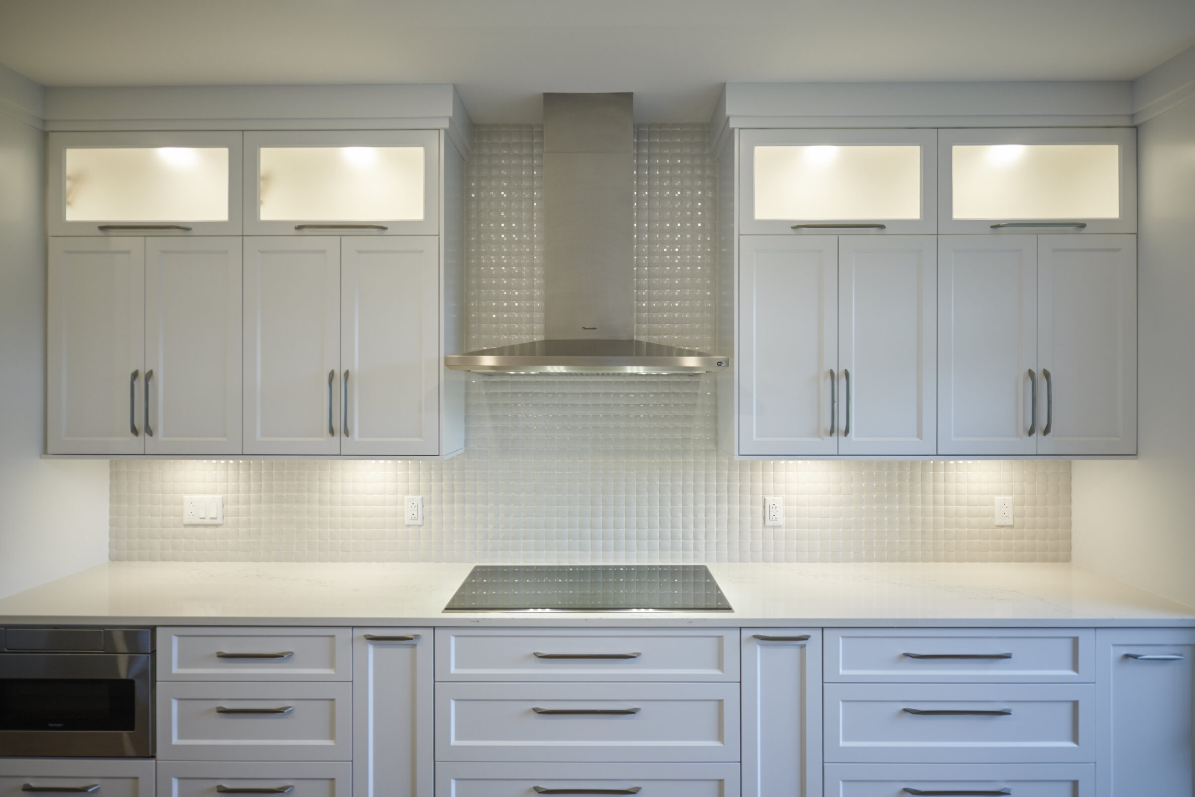 This image shows a modern kitchen with light gray cabinets, stainless steel range hood, white countertops, a cooktop, and under-cabinet lighting.