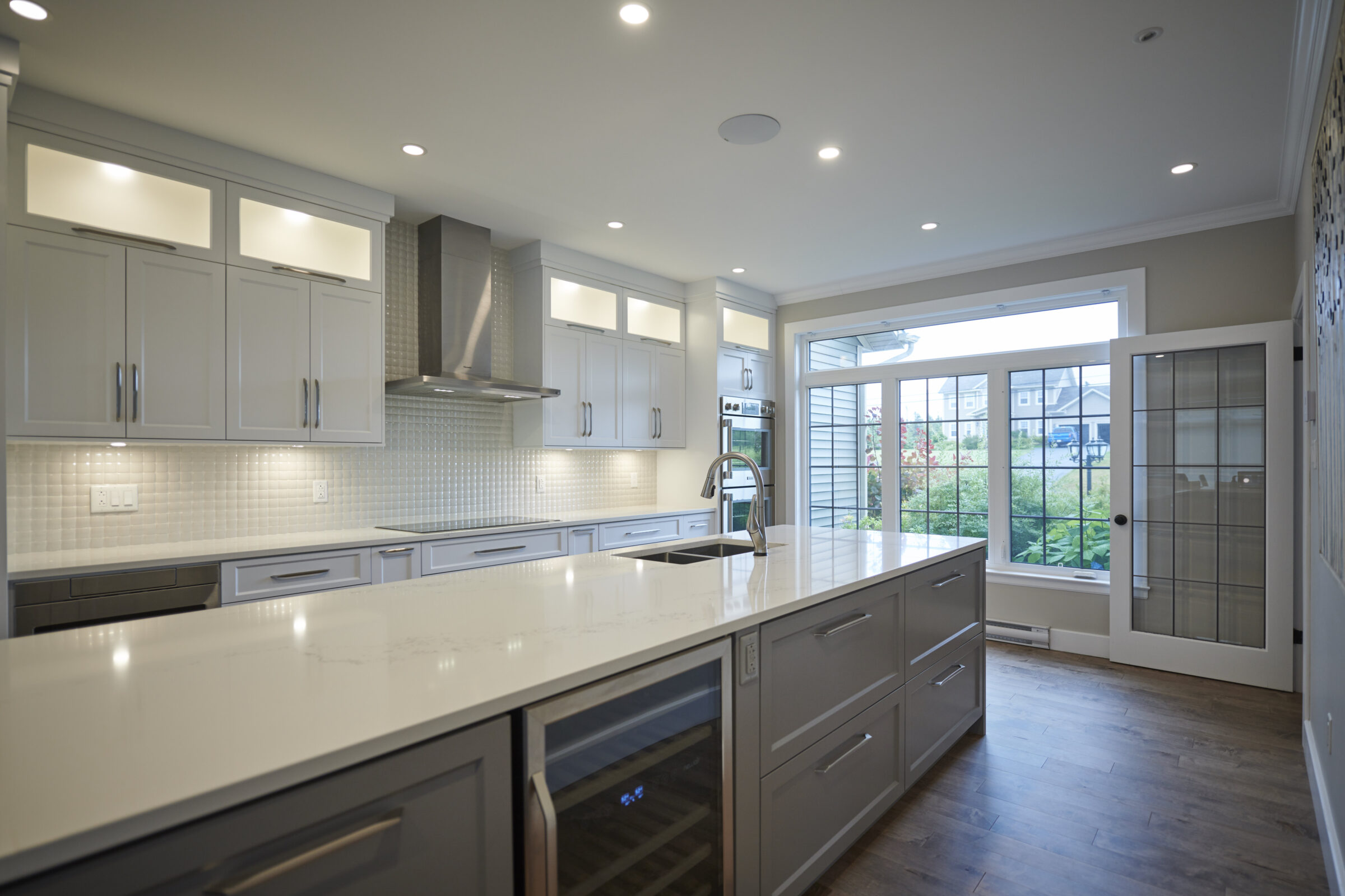 Modern kitchen interior with white cabinetry, stainless steel appliances, recessed lighting, and a view to the outdoors through large windows and glass door.