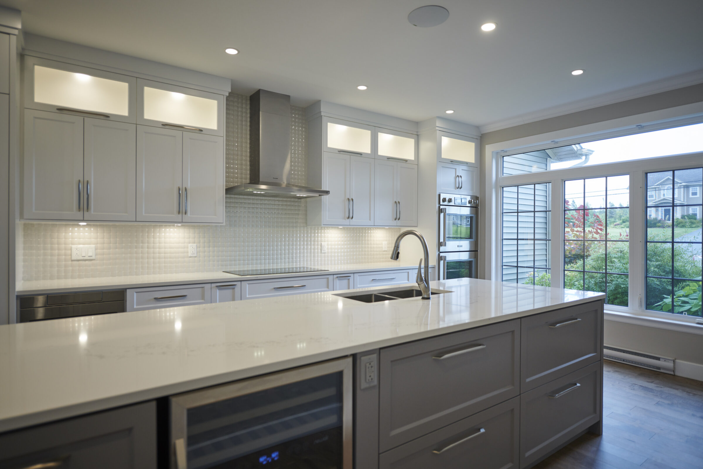 This is a modern kitchen with white cabinetry, stainless steel appliances, a large island with a sink, and a view of the outside through large windows.