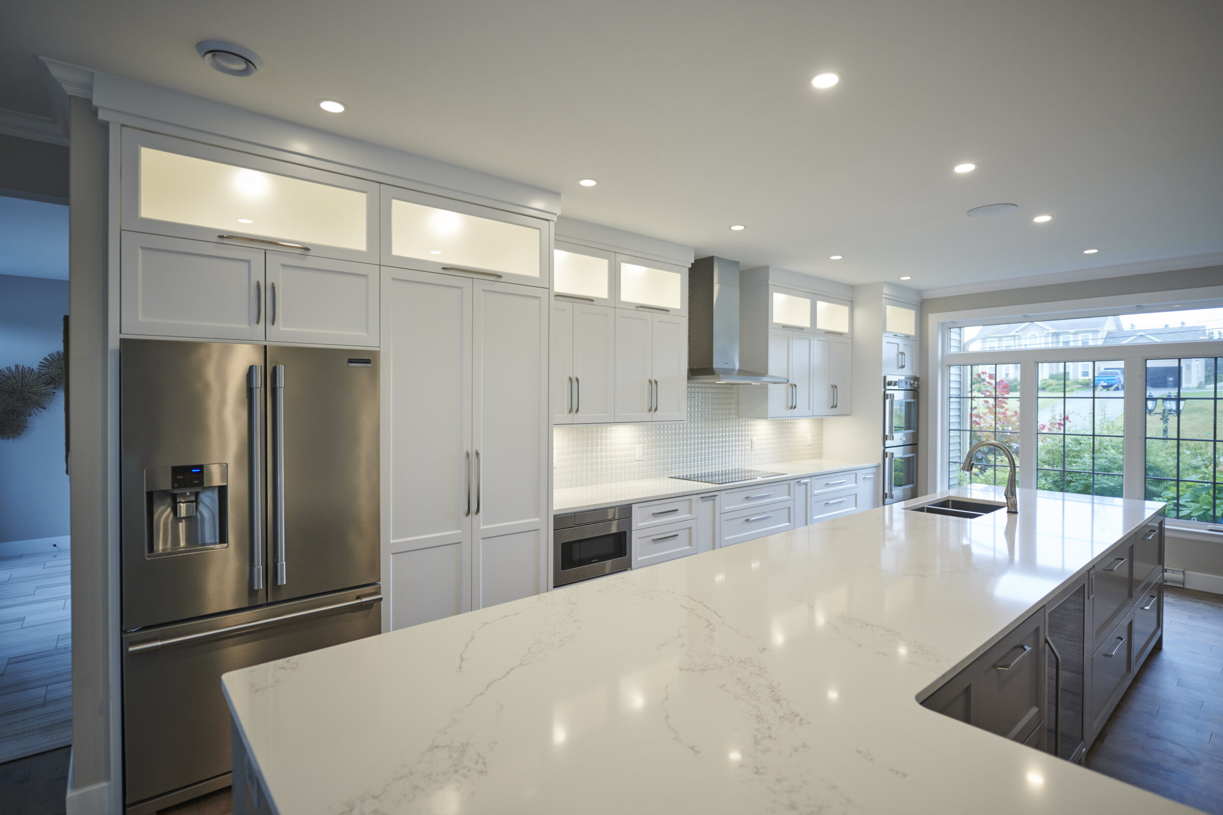 Modern kitchen with white cabinetry, marble countertops, stainless steel refrigerator, and hardwood flooring, featuring recessed lighting and a garden view.