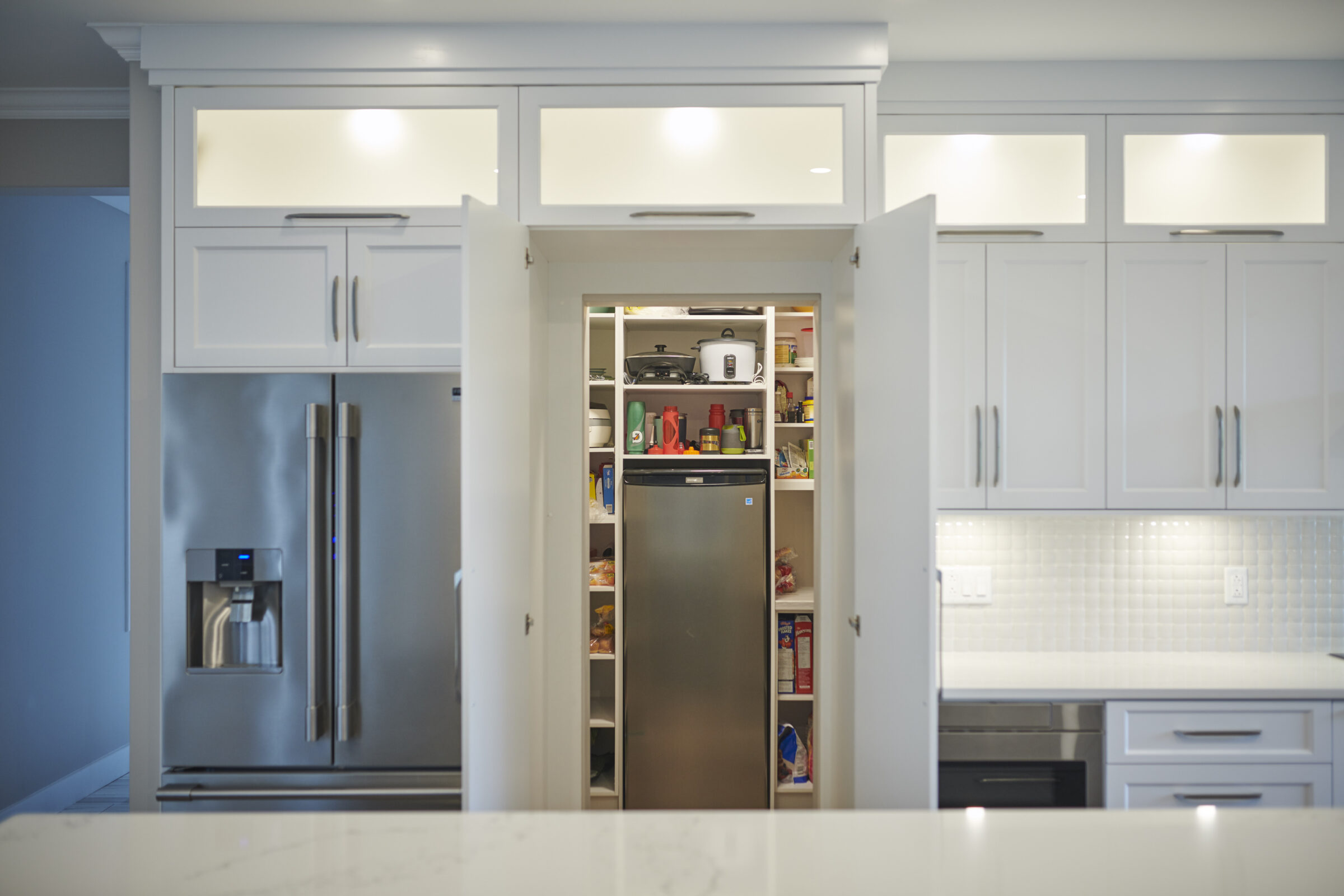 A modern kitchen with white cabinetry and stainless steel appliances, including a refrigerator and a pantry door open revealing shelves stocked with various items.