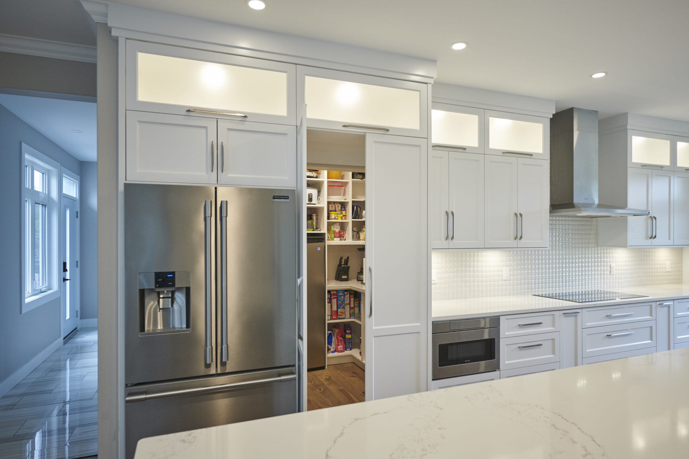 This modern kitchen features white cabinetry, stainless steel appliances, a marble countertop, and an open pantry with shelves stocked with food items.