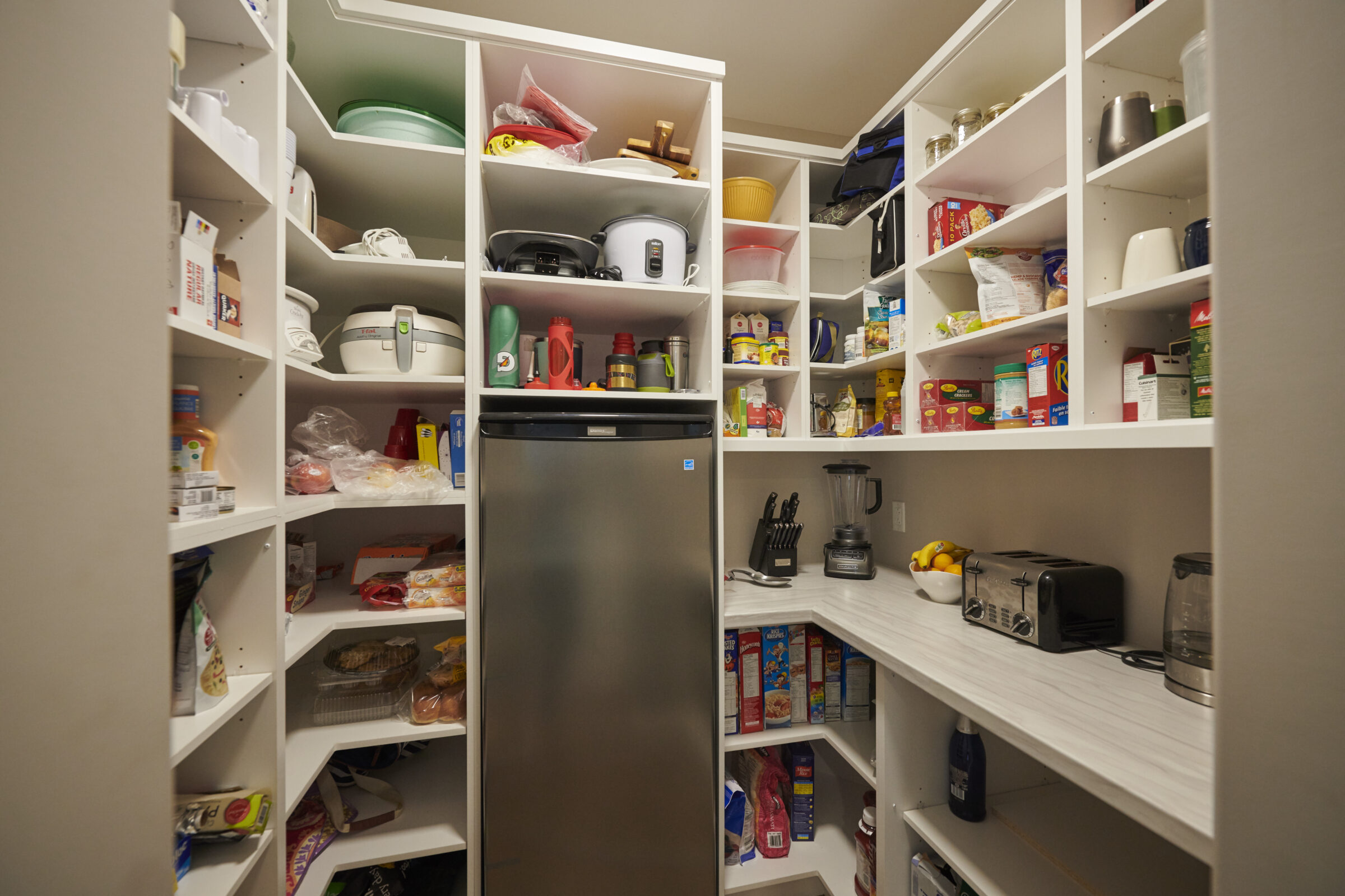This image shows a well-organized pantry with shelves stocked with various foods, kitchen appliances, and a refrigerator, suggesting a domestic setting.