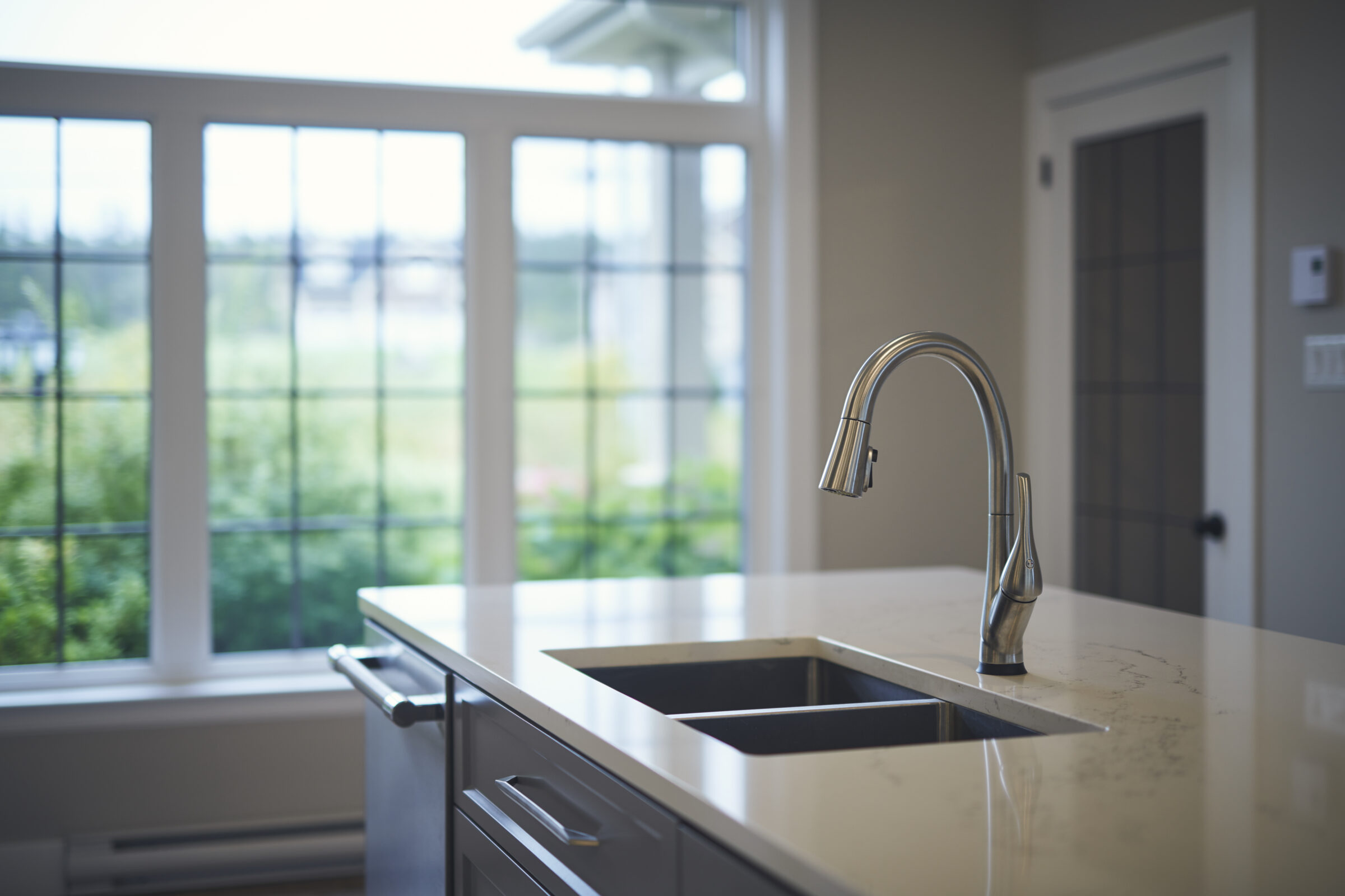 A modern kitchen with a stainless steel faucet over a sink, marble countertop, and spacious windows allowing natural light overlooking a green landscape.