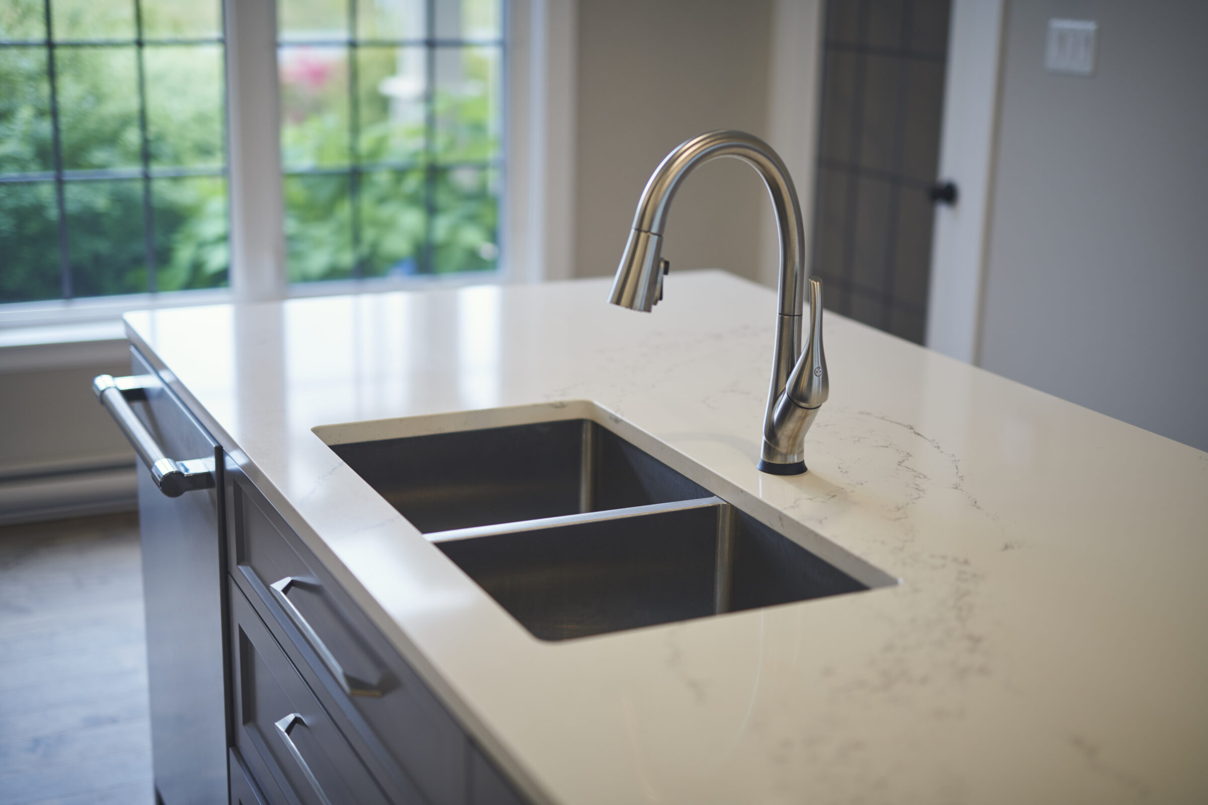 A modern kitchen countertop with a stainless steel undermount sink and high-arc faucet. A window provides a view of greenery outside.