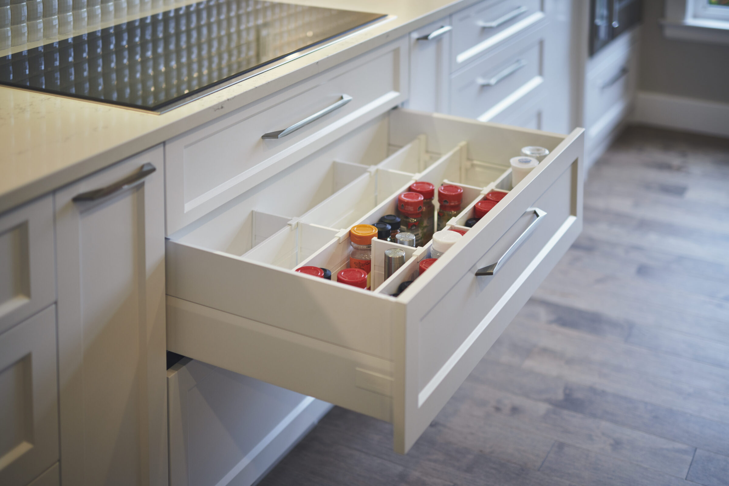 A modern kitchen drawer is open, showing an organized arrangement of spices in various jars within divided compartments. Wooden flooring complements the white cabinetry.