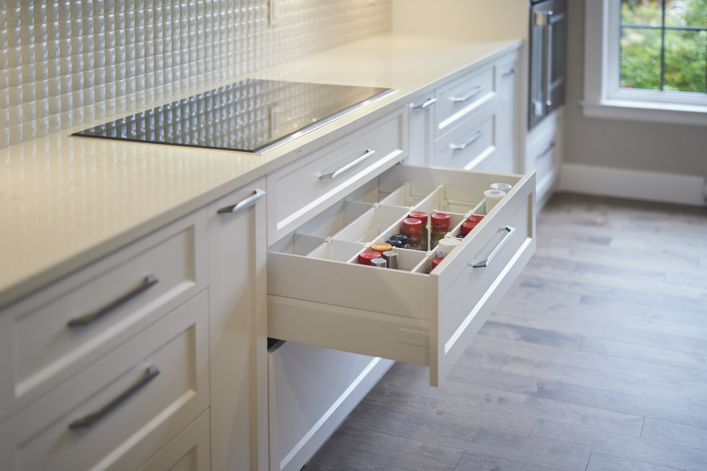 A modern kitchen with an open drawer revealing organized spices. Stainless steel cooktop, pale backsplash, and wood flooring complement the cream cabinets.