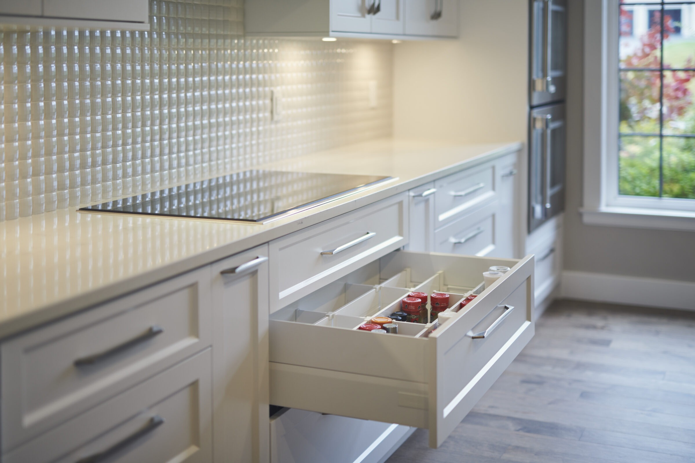 A modern kitchen with white cabinetry, a textured backsplash, and open drawers displaying organized spices. Natural light from a window enhances the space.