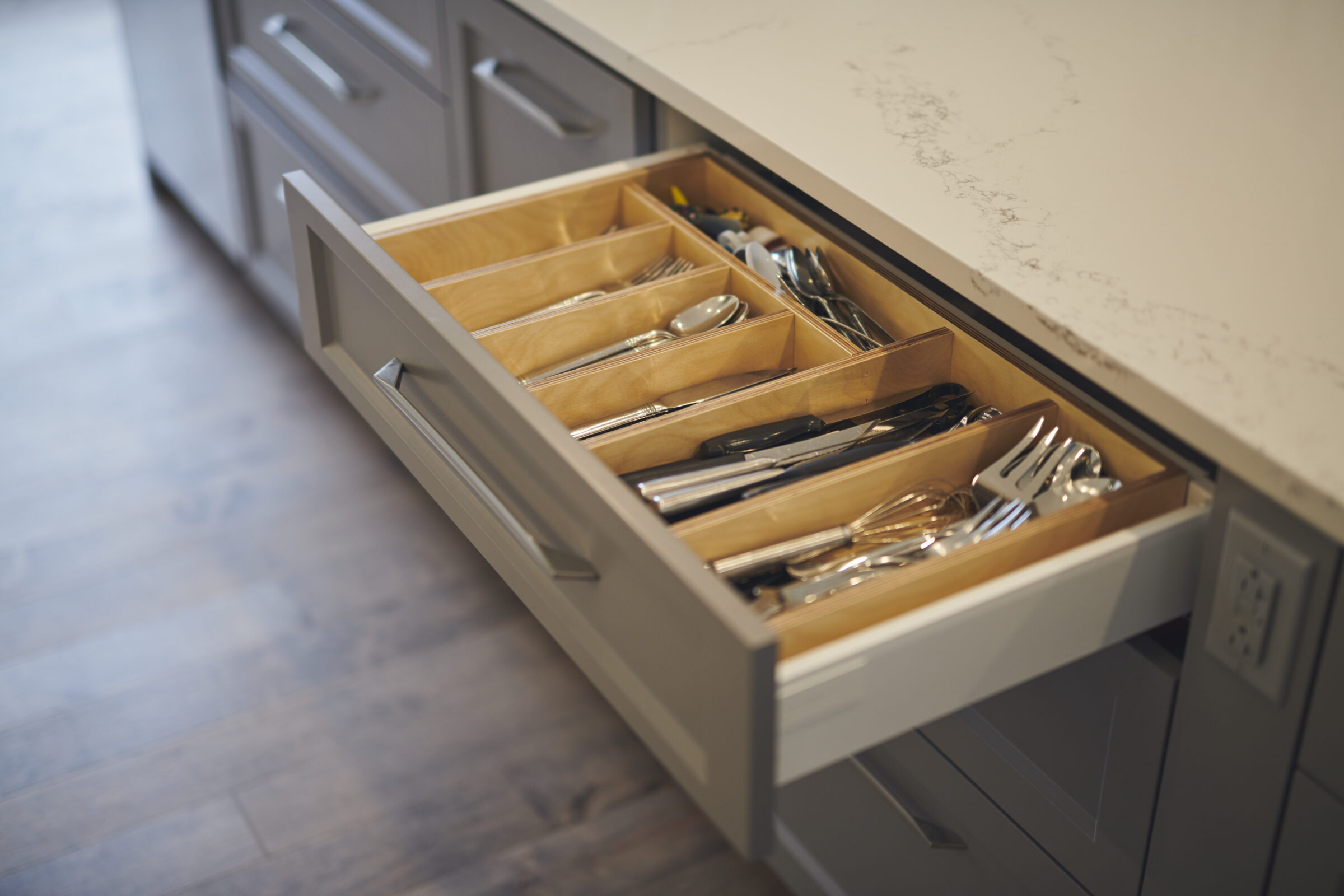 An open kitchen drawer displaying neatly arranged utensils including forks, knives, and spoons, with a wooden organizer in a modern kitchen setting.