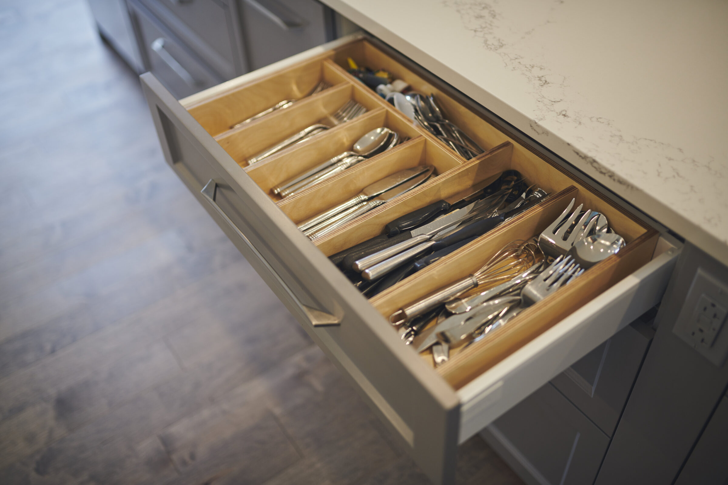 An open kitchen drawer displaying an organized assortment of cutlery, including spoons, forks, knives, and utensils, with a wooden organizer in a modern setting.