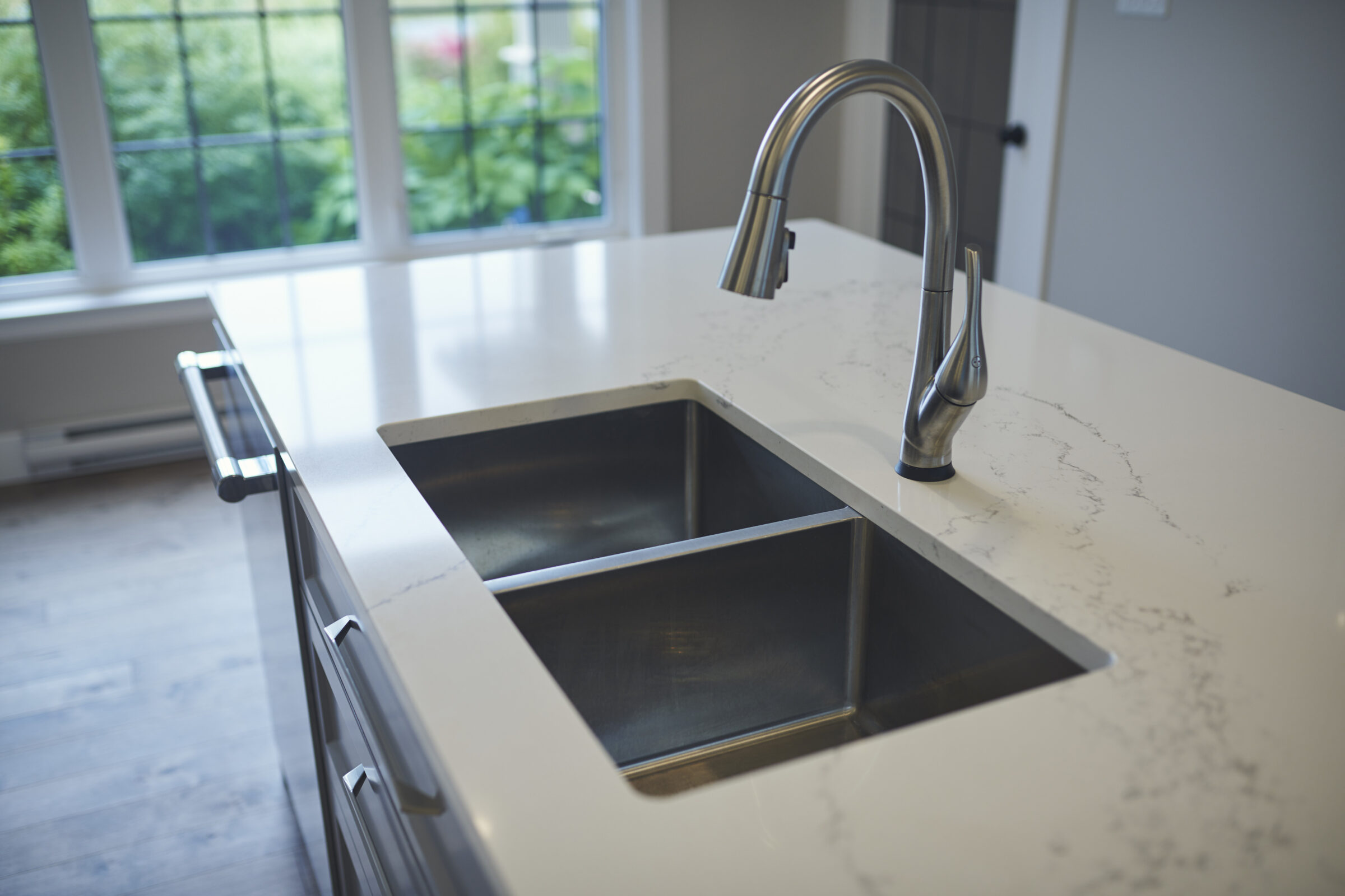 This image shows a modern kitchen with a stainless steel double sink, marble countertop, and a large window with a view of greenery outside.