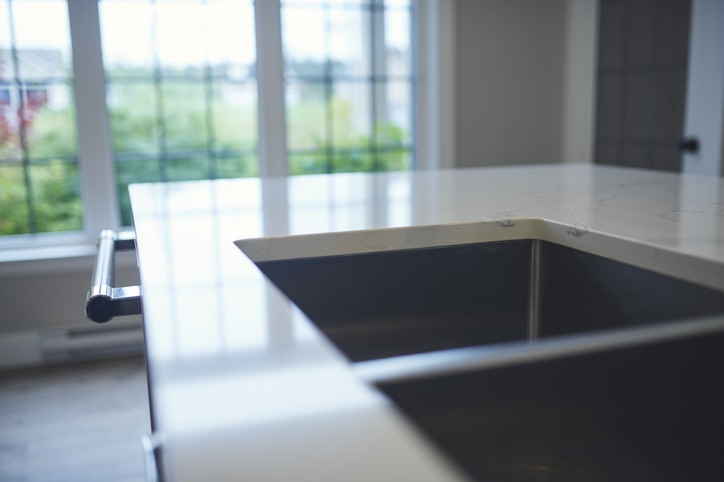 A modern kitchen interior with a close-up of a clean black undermount sink, stainless steel faucet, white countertop, and a window overlooking outdoor greenery.