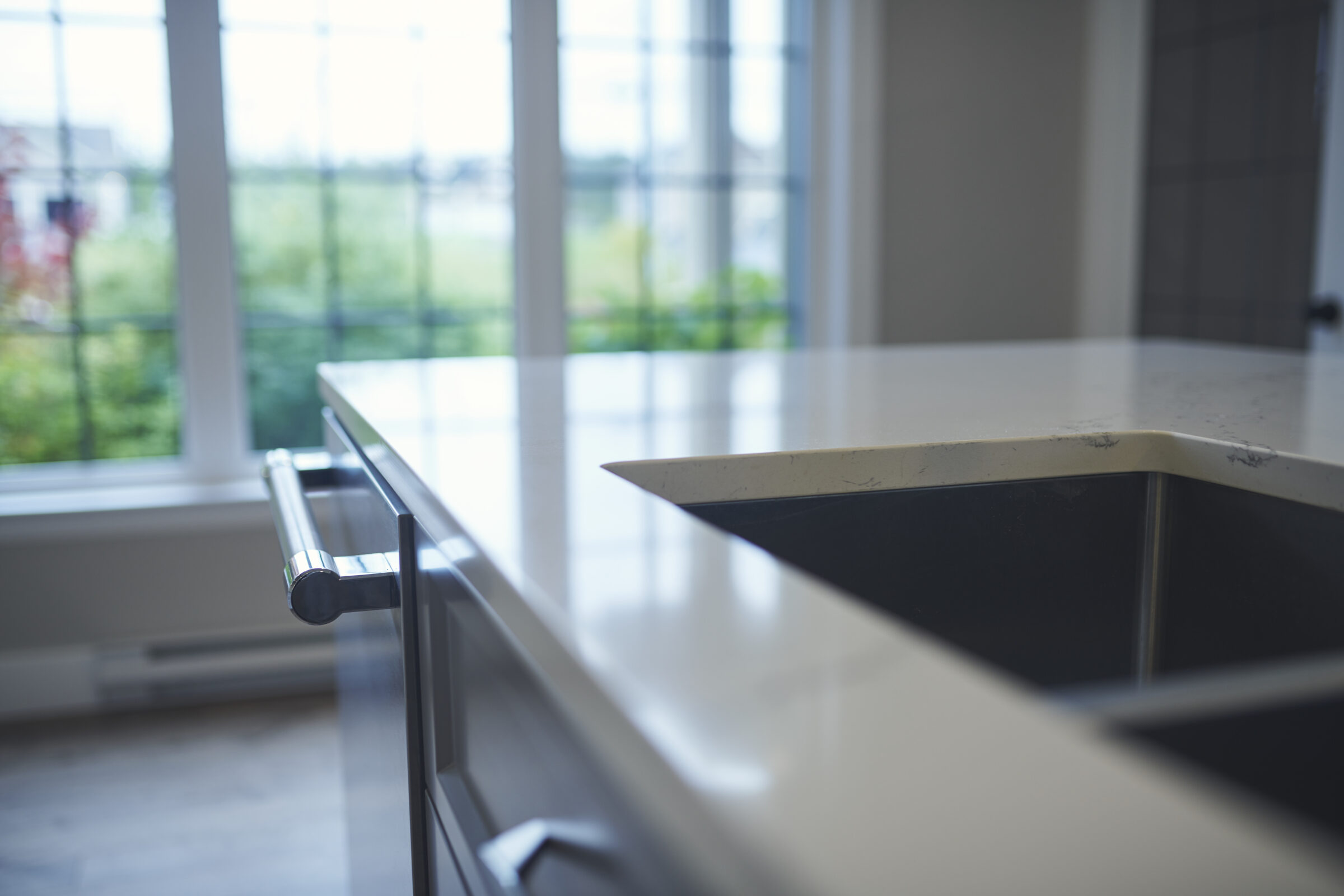 Modern kitchen counter with a built-in cooktop, sleek handle, and reflective surface, overlooking a window with a view of greenery outside.