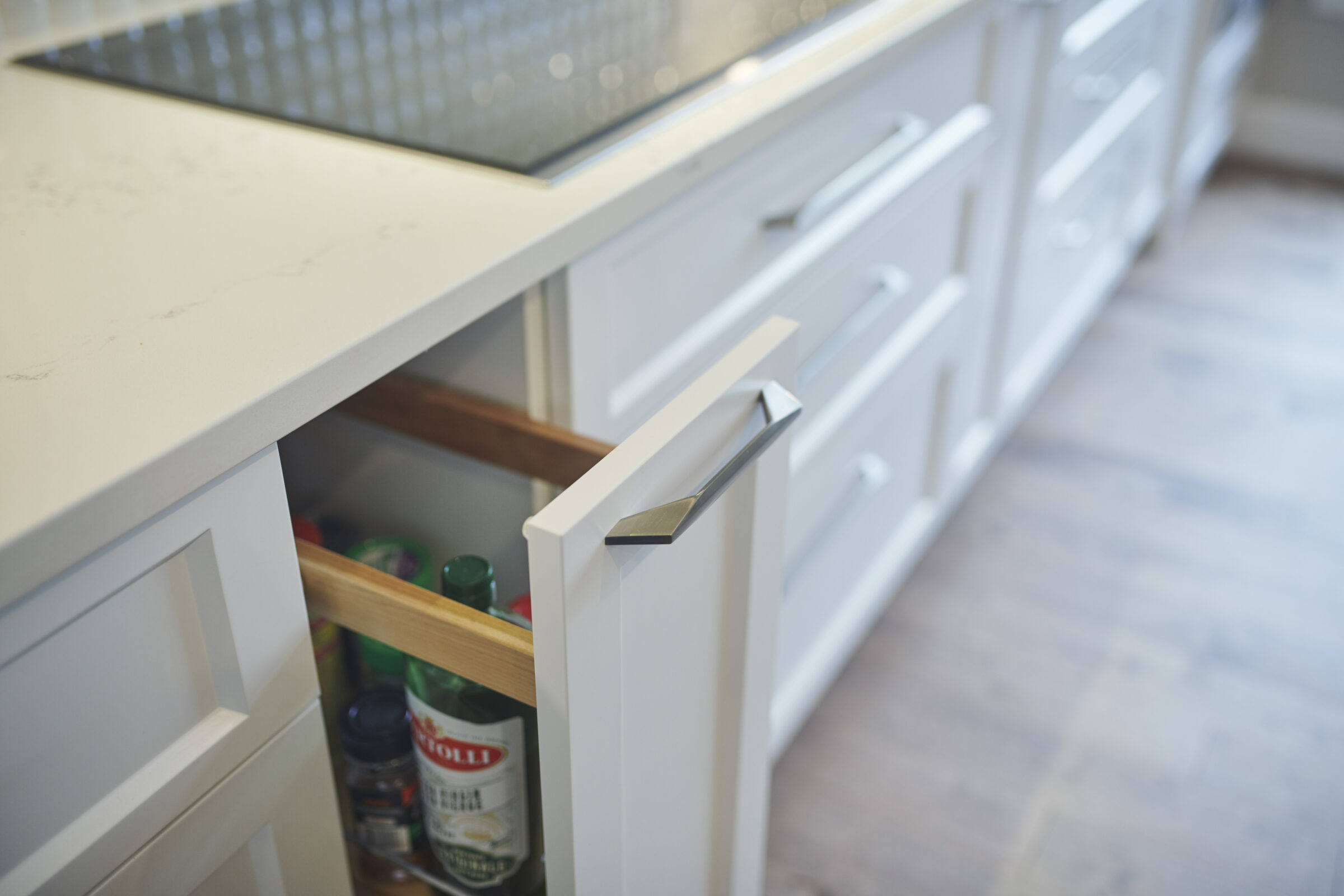 A modern kitchen drawer is slightly open, revealing bottles of condiments inside. The focus is on the sleek metal handle against white cabinetry.