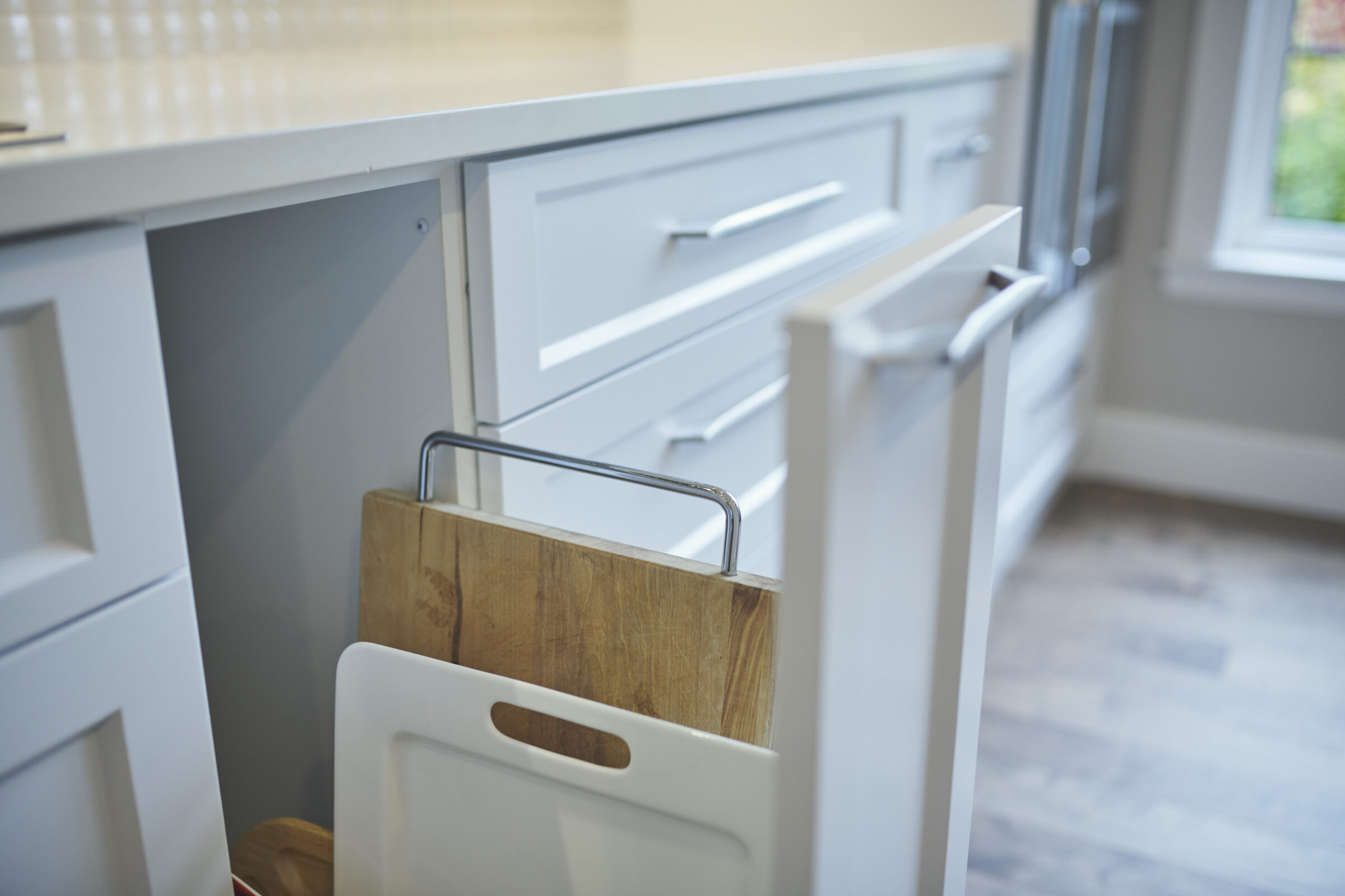A modern kitchen with white cabinetry and stainless steel handles, featuring a wooden cutting board stored in a pull-out rack, with soft natural light.