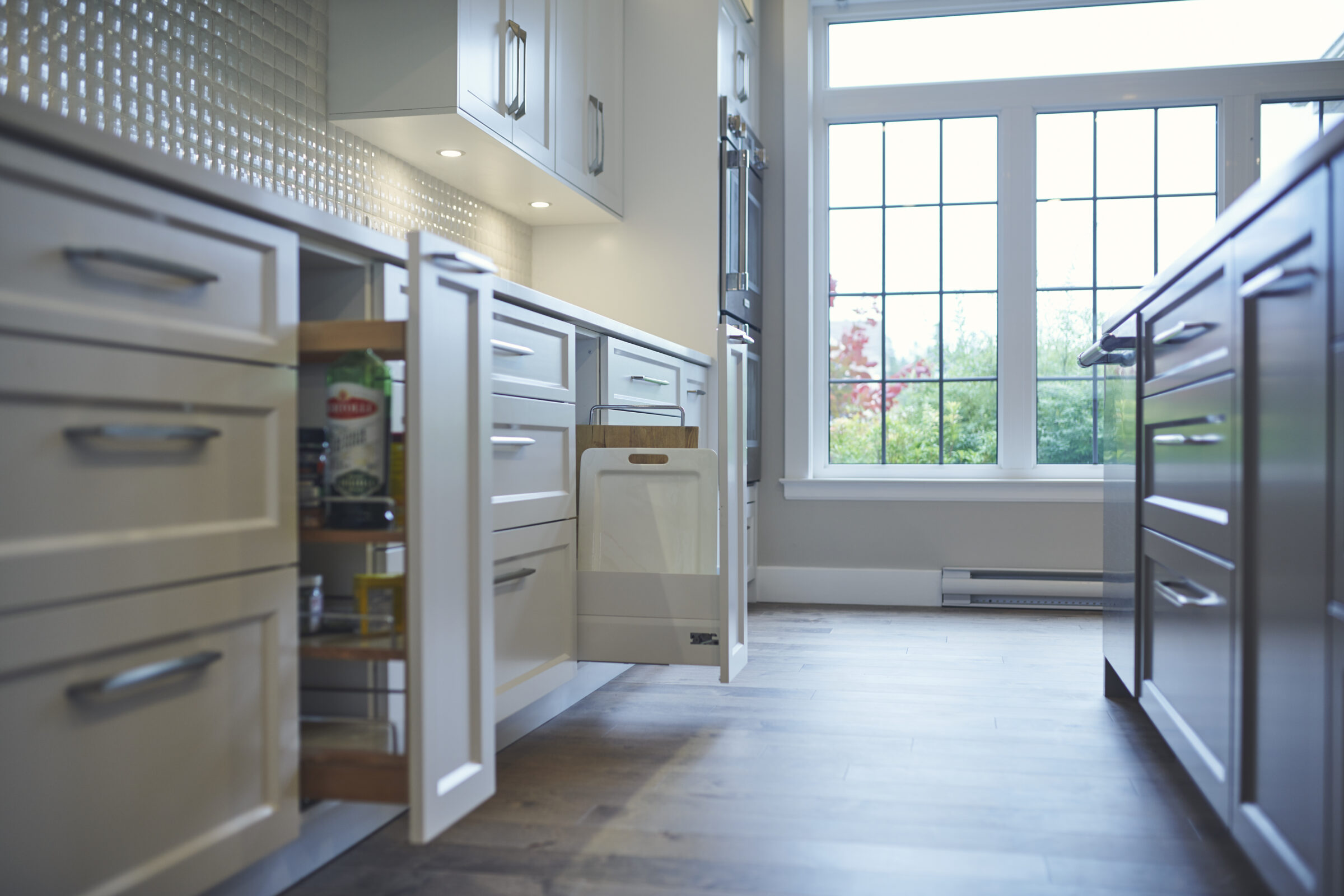 A modern kitchen with white cabinetry, stainless steel handles, and a large window providing natural light. Drawers are pulled open, revealing storage space.