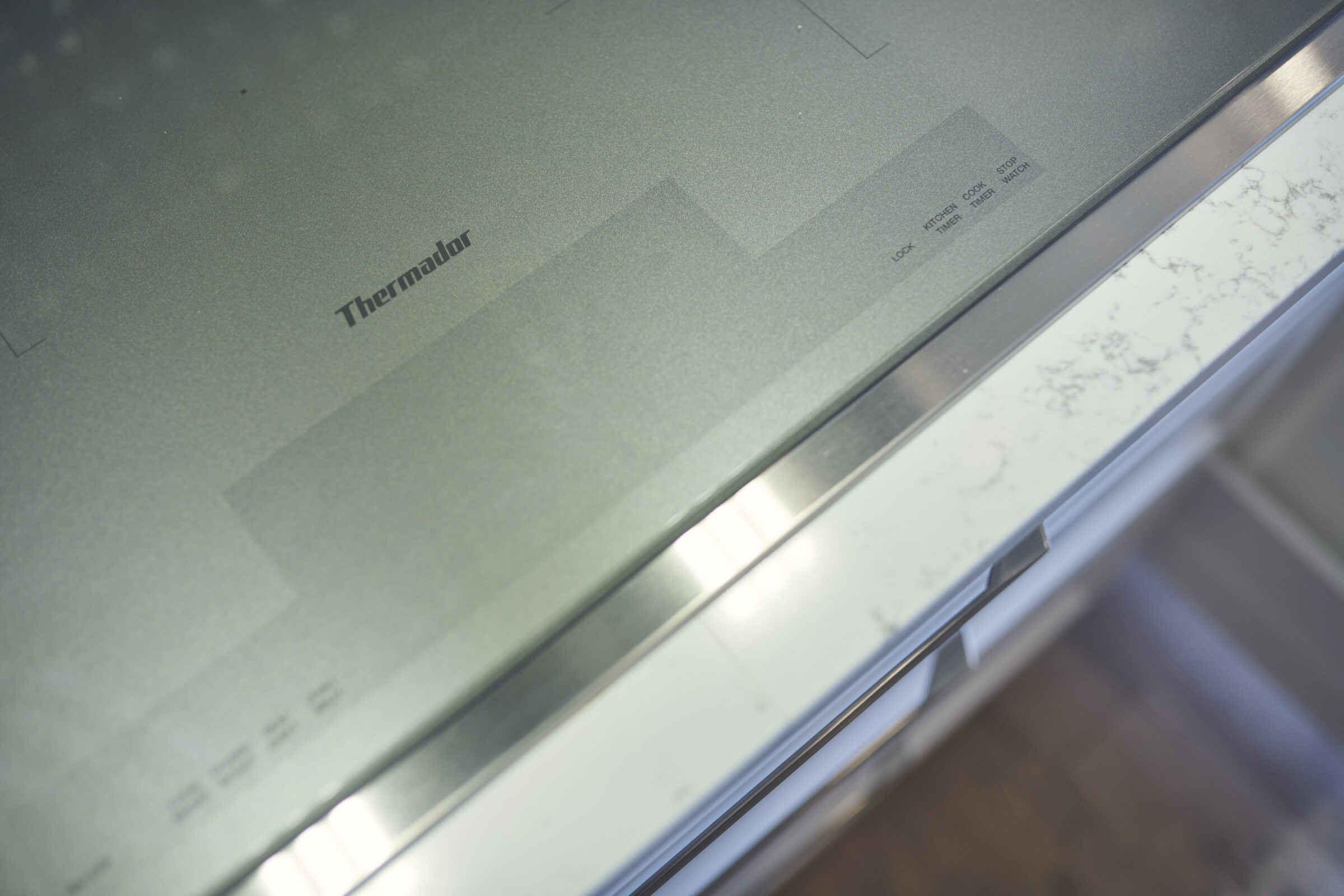 The image shows a close-up view of a modern, stainless steel Thermador cooking appliance with control panel text, situated above marble countertops in a kitchen.