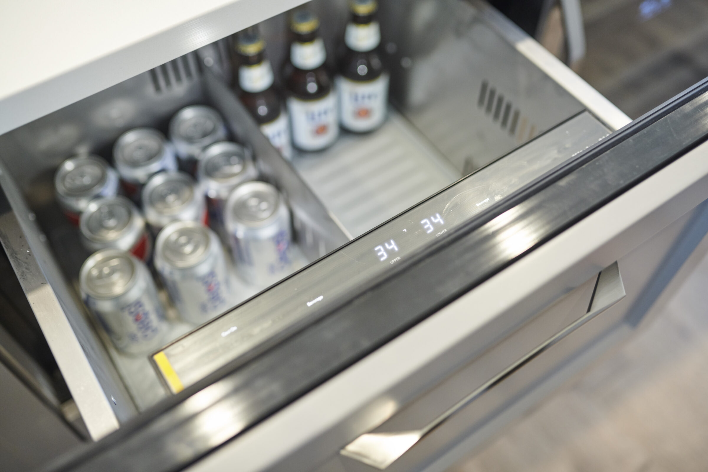 An open refrigerator drawer displays cans and bottles with a temperature reading of 34 degrees, likely indicating a chilled setting for beverages.