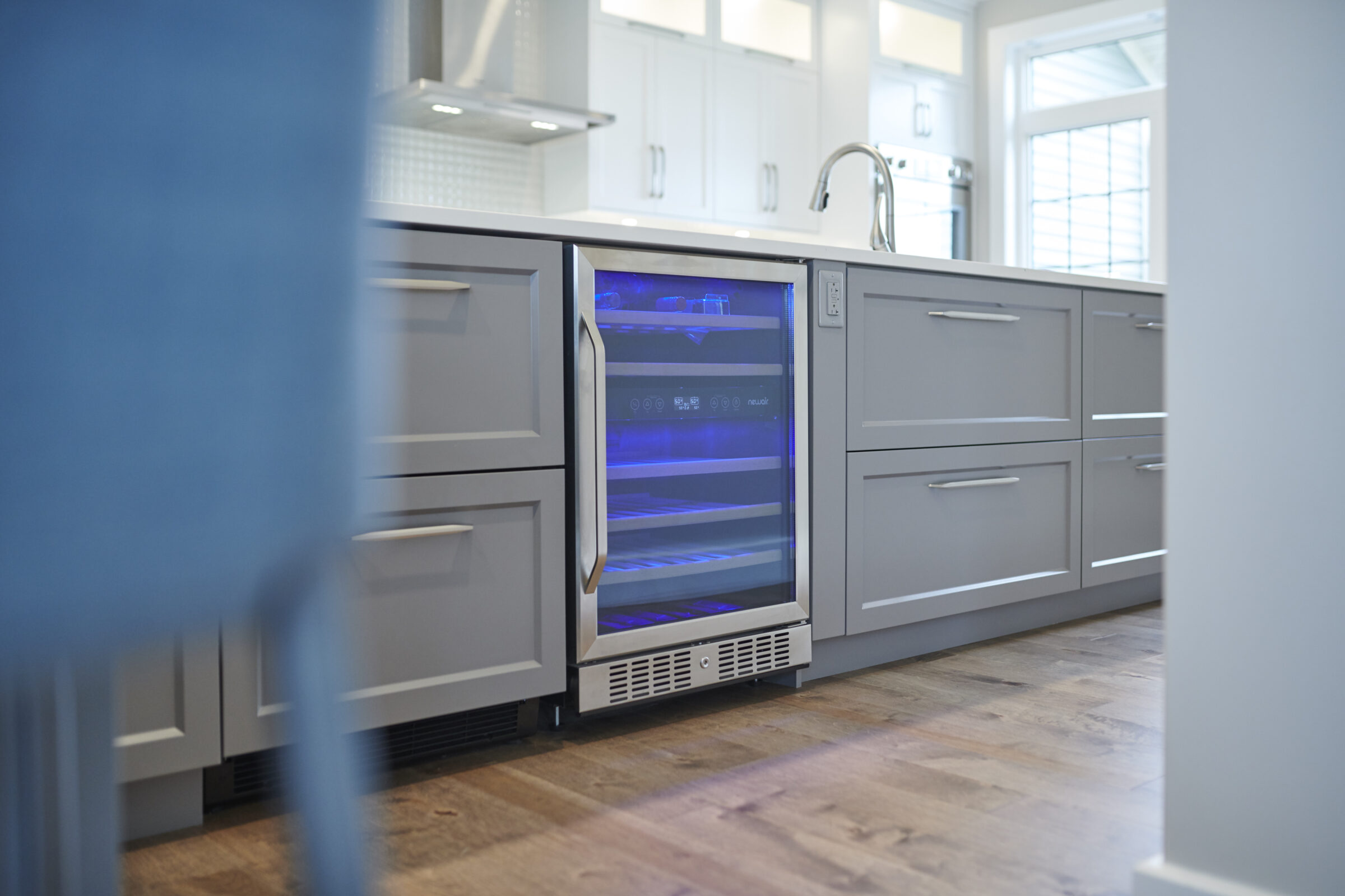 A modern kitchen with grey cabinets, a built-in wine cooler with blue lighting, wooden floors, and stainless-steel appliances visible in the background.
