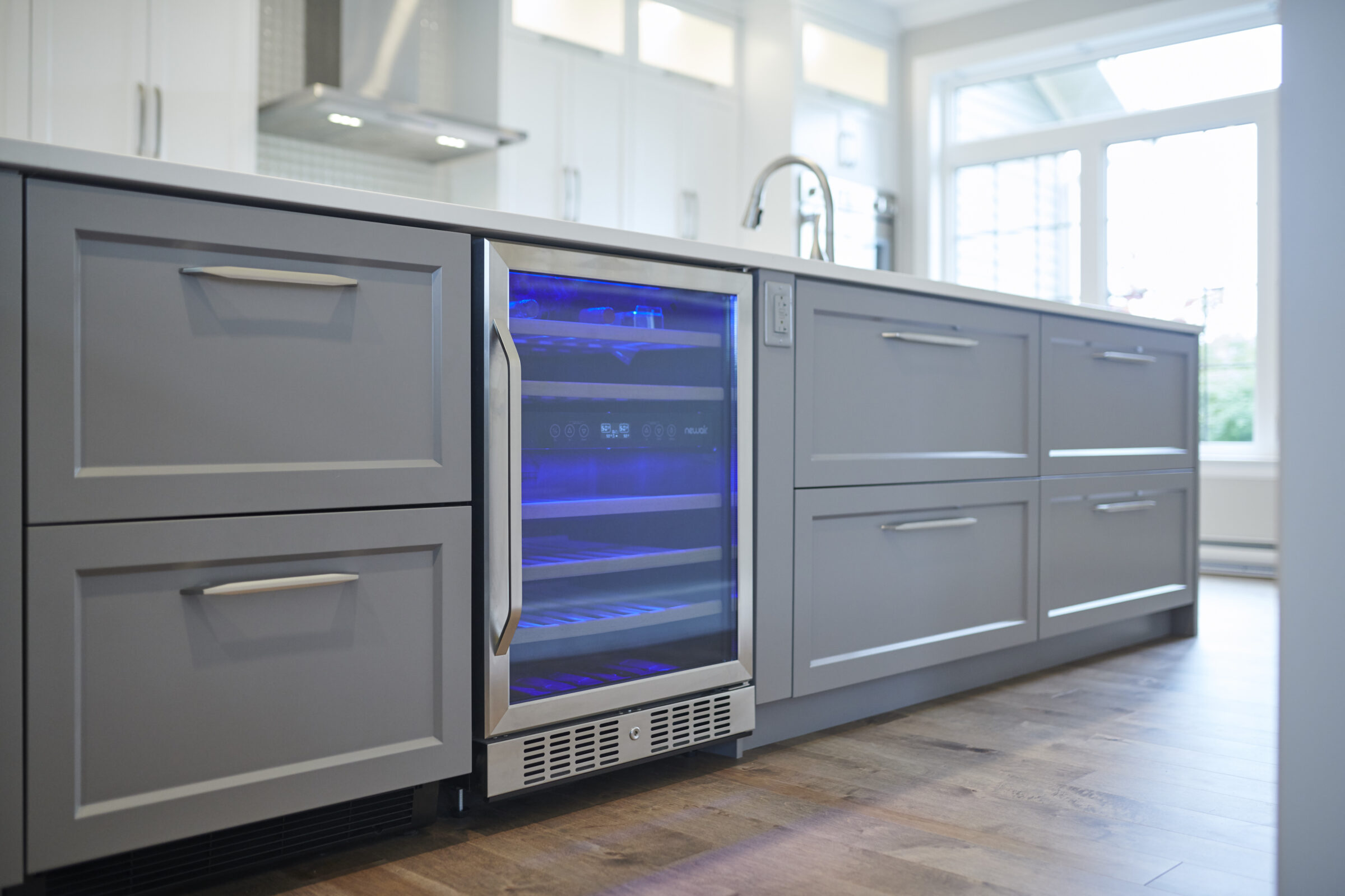 Modern kitchen interior with grey cabinets and a built-in wine cooler with a glass door illuminated by blue light. Hardwood flooring and a window are visible.