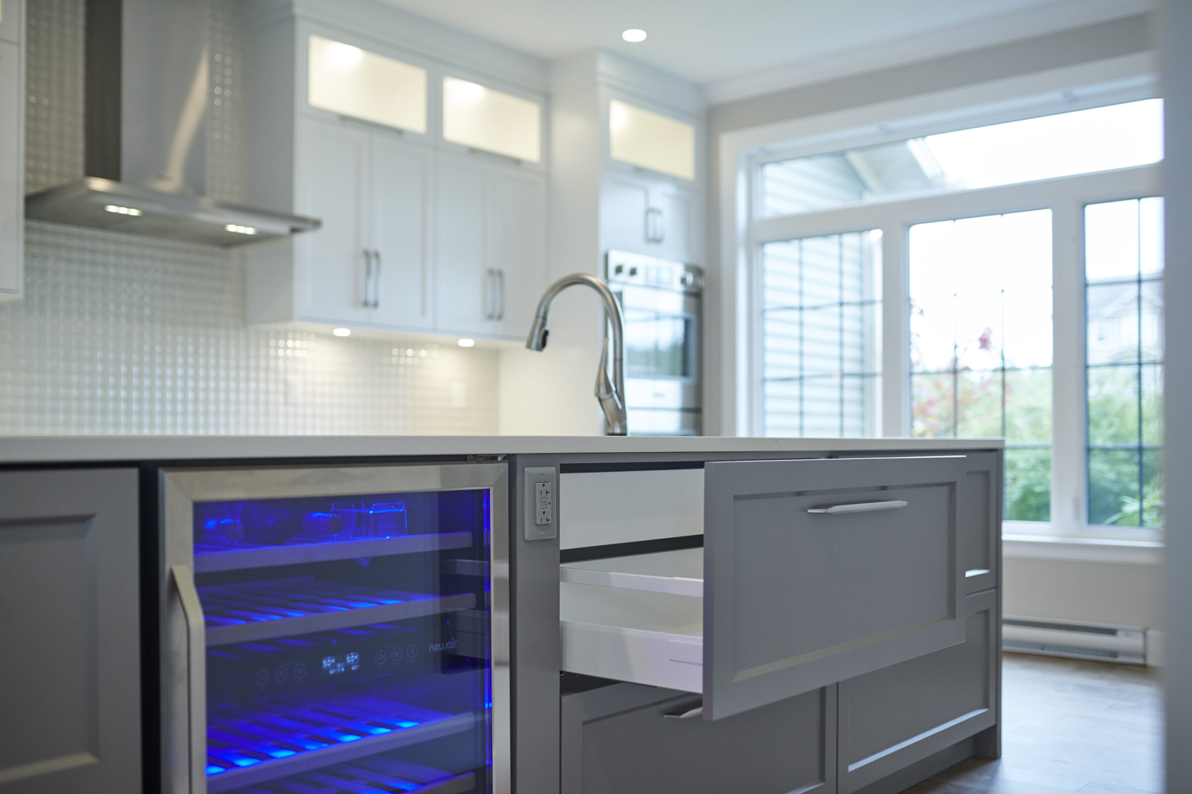 A modern kitchen with white cabinetry and stainless steel appliances. A wine fridge glows blue, and a large window lets in natural light.