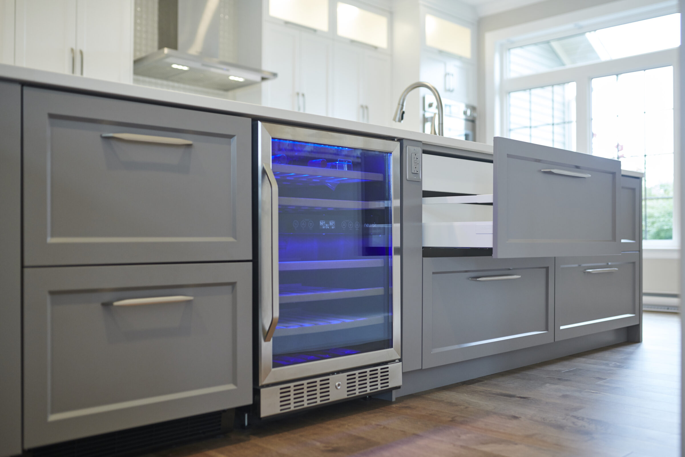 A modern kitchen with gray cabinets, a wine cooler with blue lighting, stainless steel handles, white countertops, and hardwood floors.
