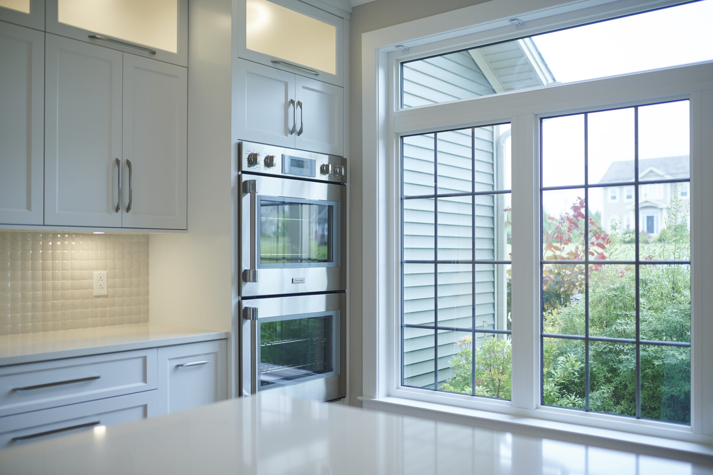 A modern kitchen with white cabinetry, stainless steel double oven, tile backsplash, and large windows offering a view of the outdoors.