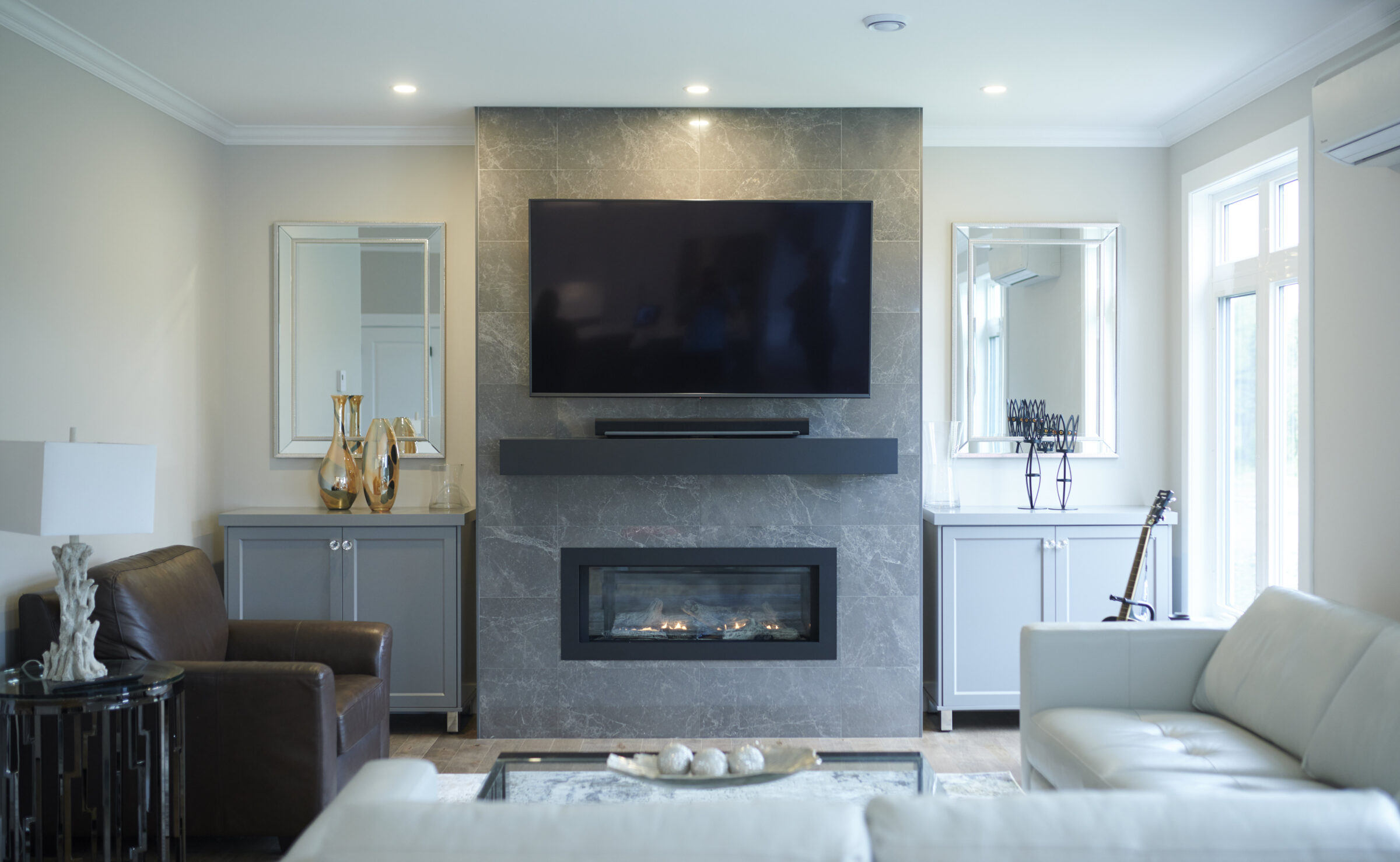 A modern living room with a stone fireplace, wall-mounted TV, leather armchair, sofa, decorations, and a guitar resting against a cabinet near windows.